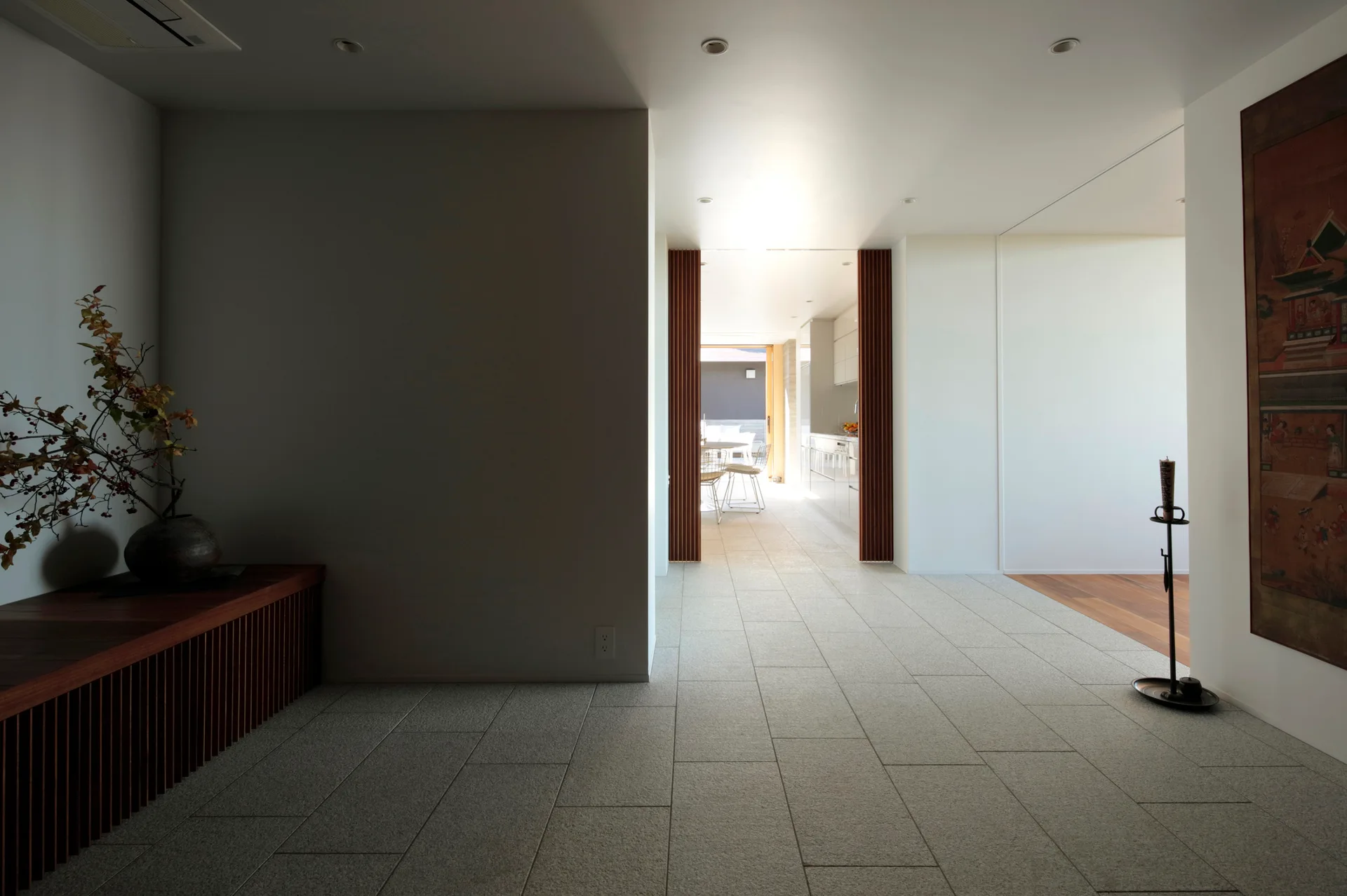A minimalist entryway features grey stone tile flooring, a slatted wood console, and a doorway framed with wooden slats leading to a bright kitchen.
