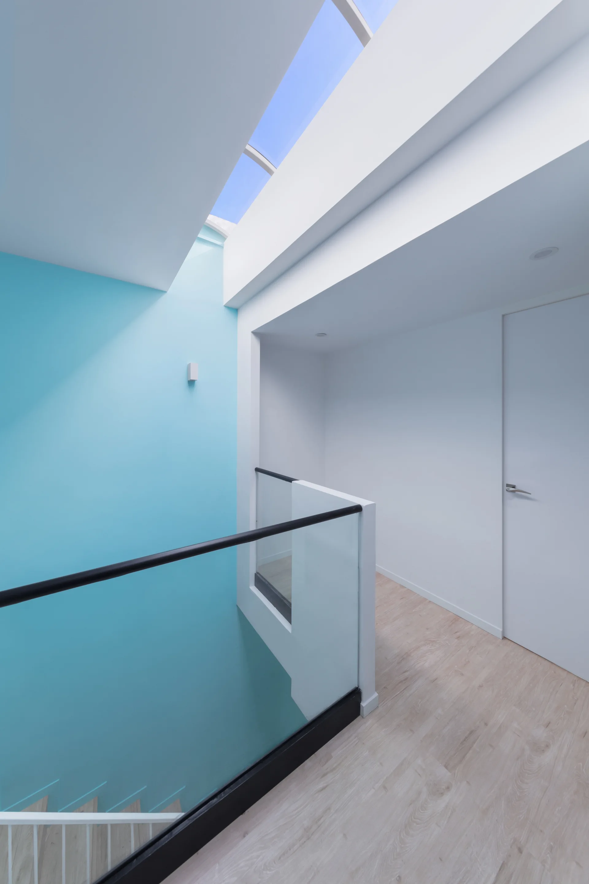 A minimalist hallway landing with a glass staircase railing, light wood floors, a bright blue accent wall, and a large skylight overhead.