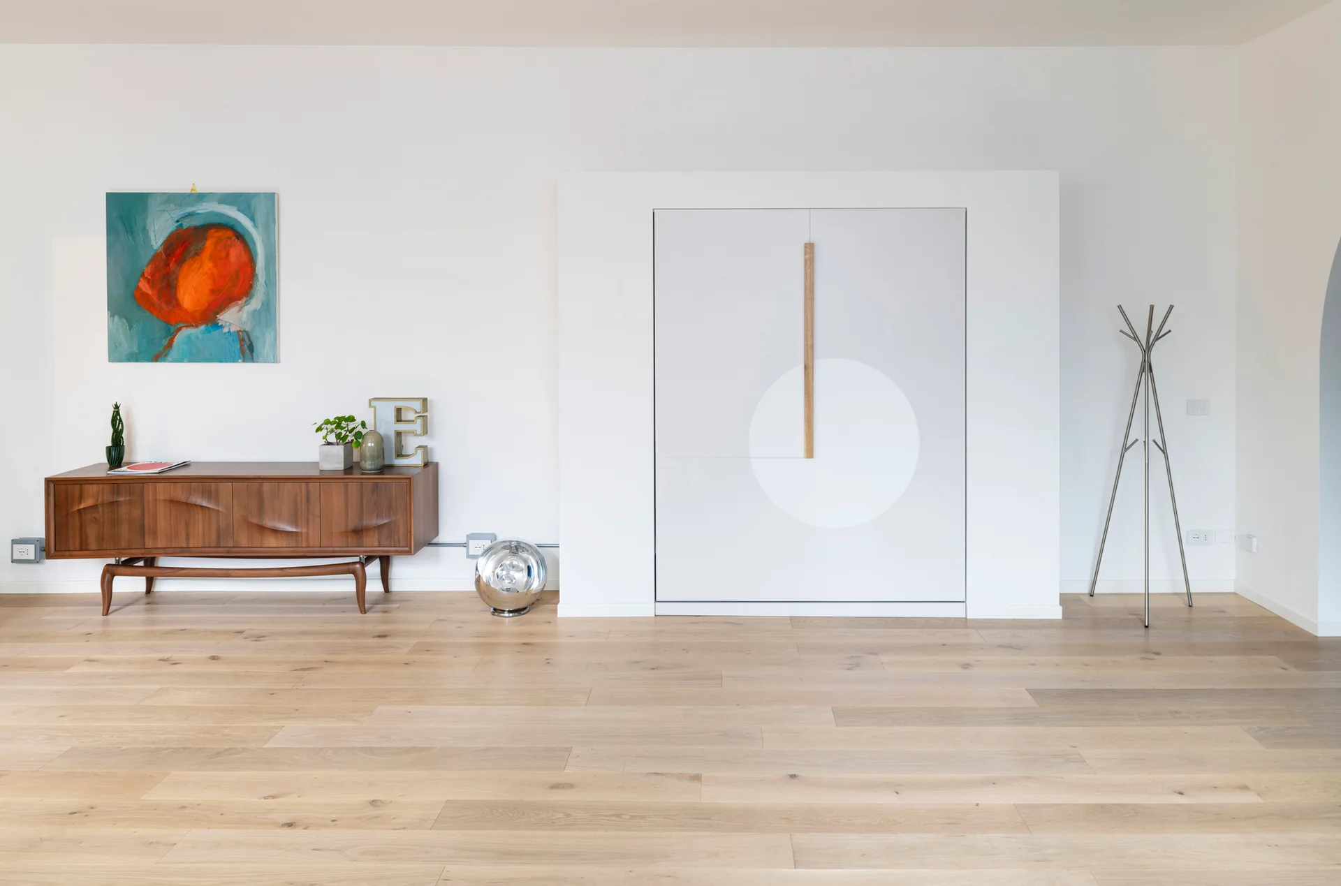 A minimalist living room features a mid-century modern wooden sideboard, a colorful abstract painting, and a large white built-in storage cabinet.
