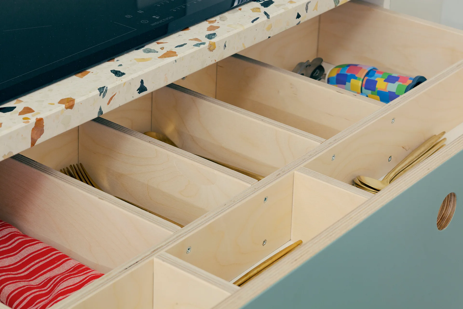 An open blue-green kitchen drawer with light wood plywood dividers holds gold cutlery beneath a speckled terrazzo countertop.