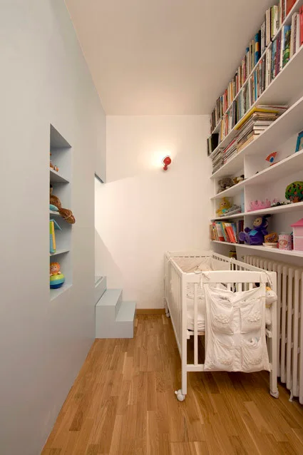 A narrow nursery features a white wooden crib, floor-to-ceiling wall-mounted bookshelves, light blue steps, and a natural wood floor.