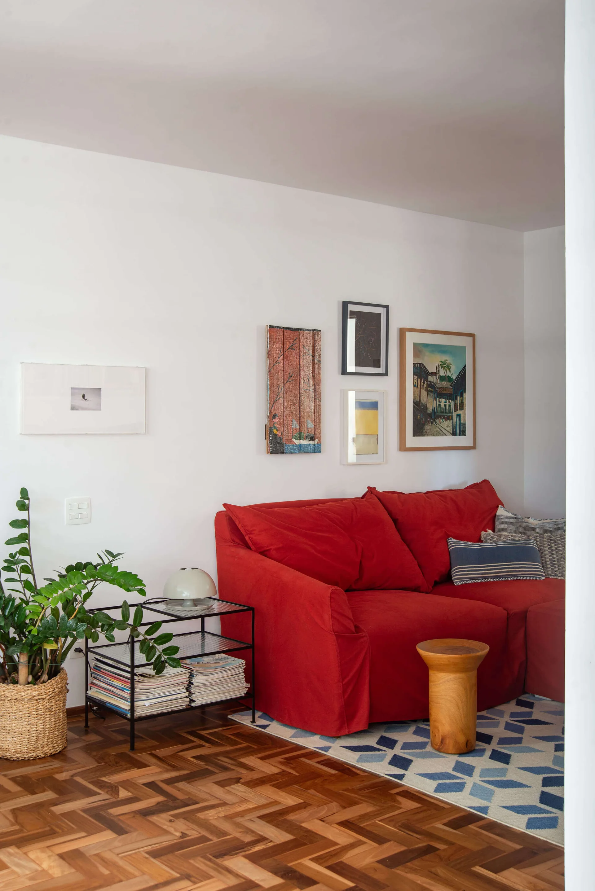 A living room features a bright red sofa set on a blue geometric rug, paired with herringbone wood flooring, a gallery wall, and a tiered side table.