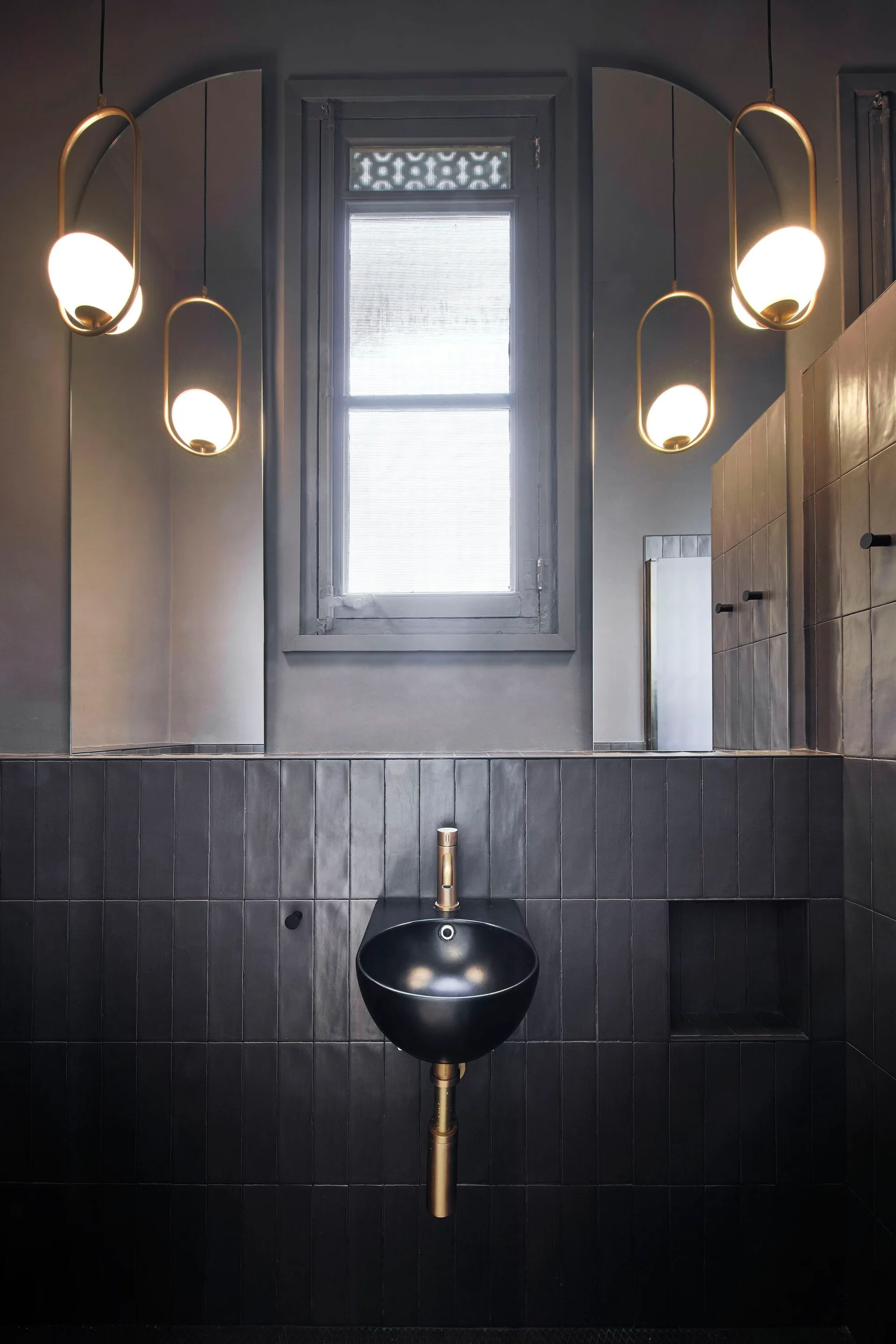 A modern bathroom vanity features a black wall-mounted sink below two arched mirrors and glowing brass globe pendant lights.