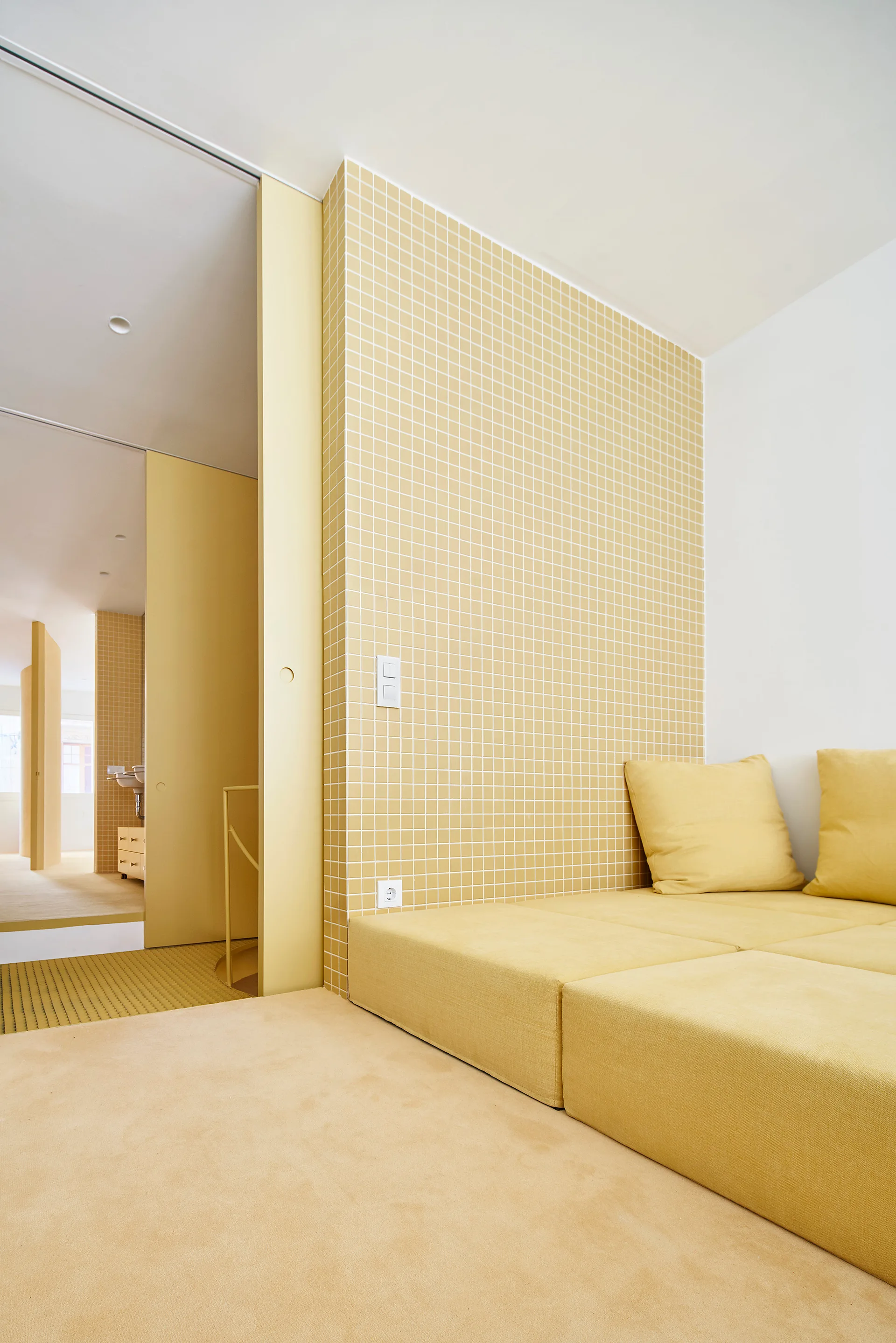 A minimalist yellow bedroom featuring a low platform bed against a yellow grid tiled accent wall with a sliding door leading to an adjacent bathroom.