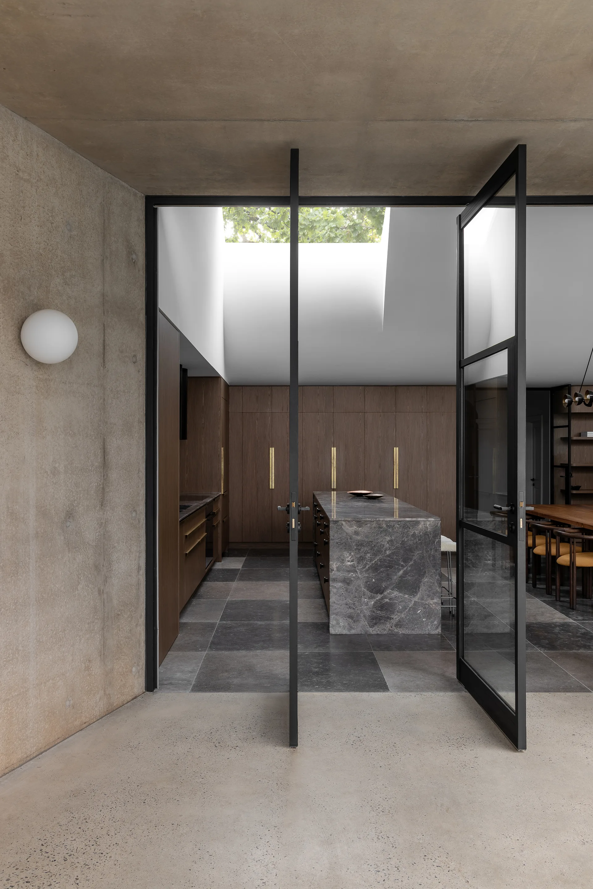 A minimalist kitchen features a dark veined marble island, wood cabinetry, a skylight, and large black-framed glass pivot doors opening from a concrete patio.