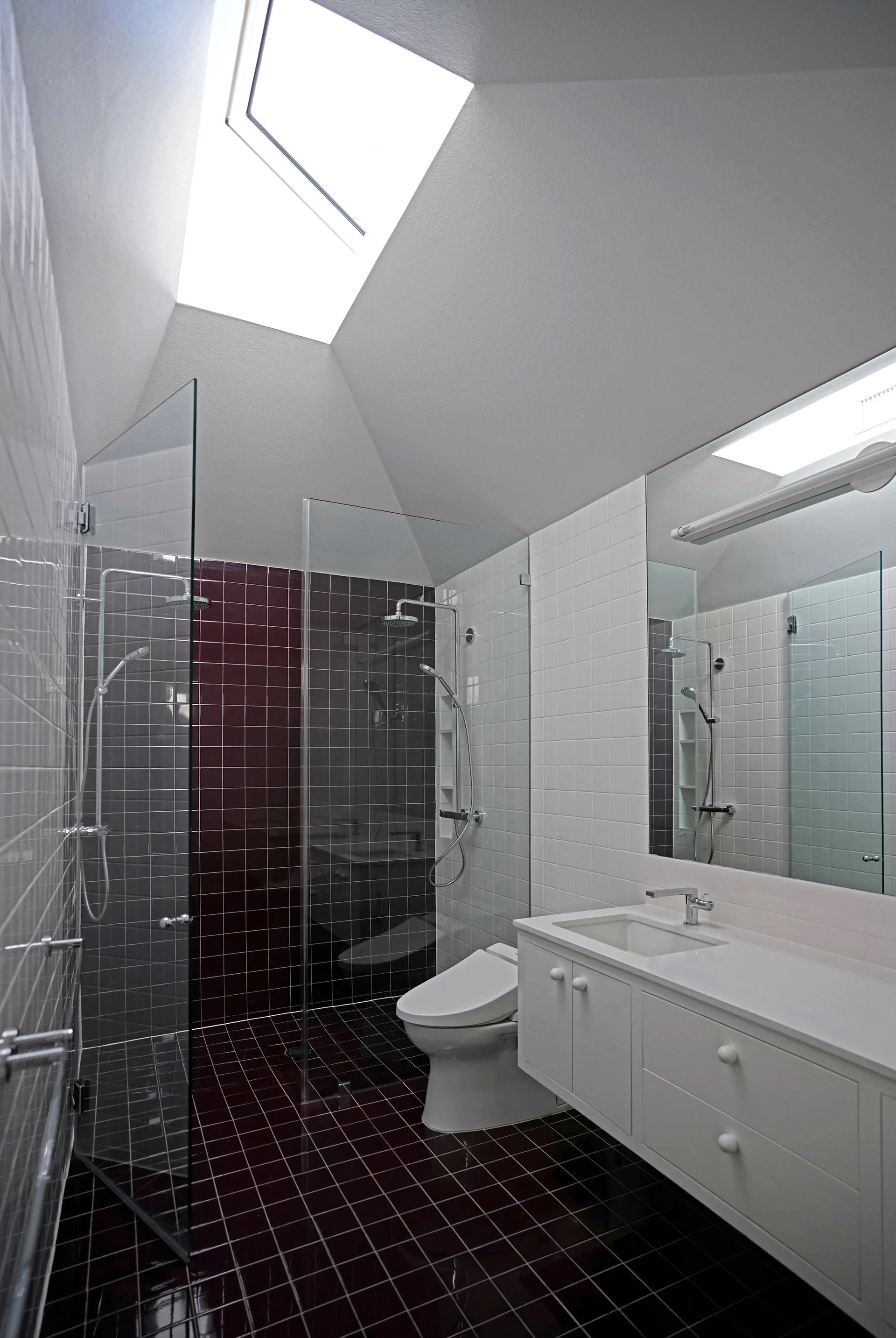 A modern bathroom featuring glossy maroon tiles, white wall tiles, a glass shower screen, a white vanity, and a skylight in the sloped ceiling.