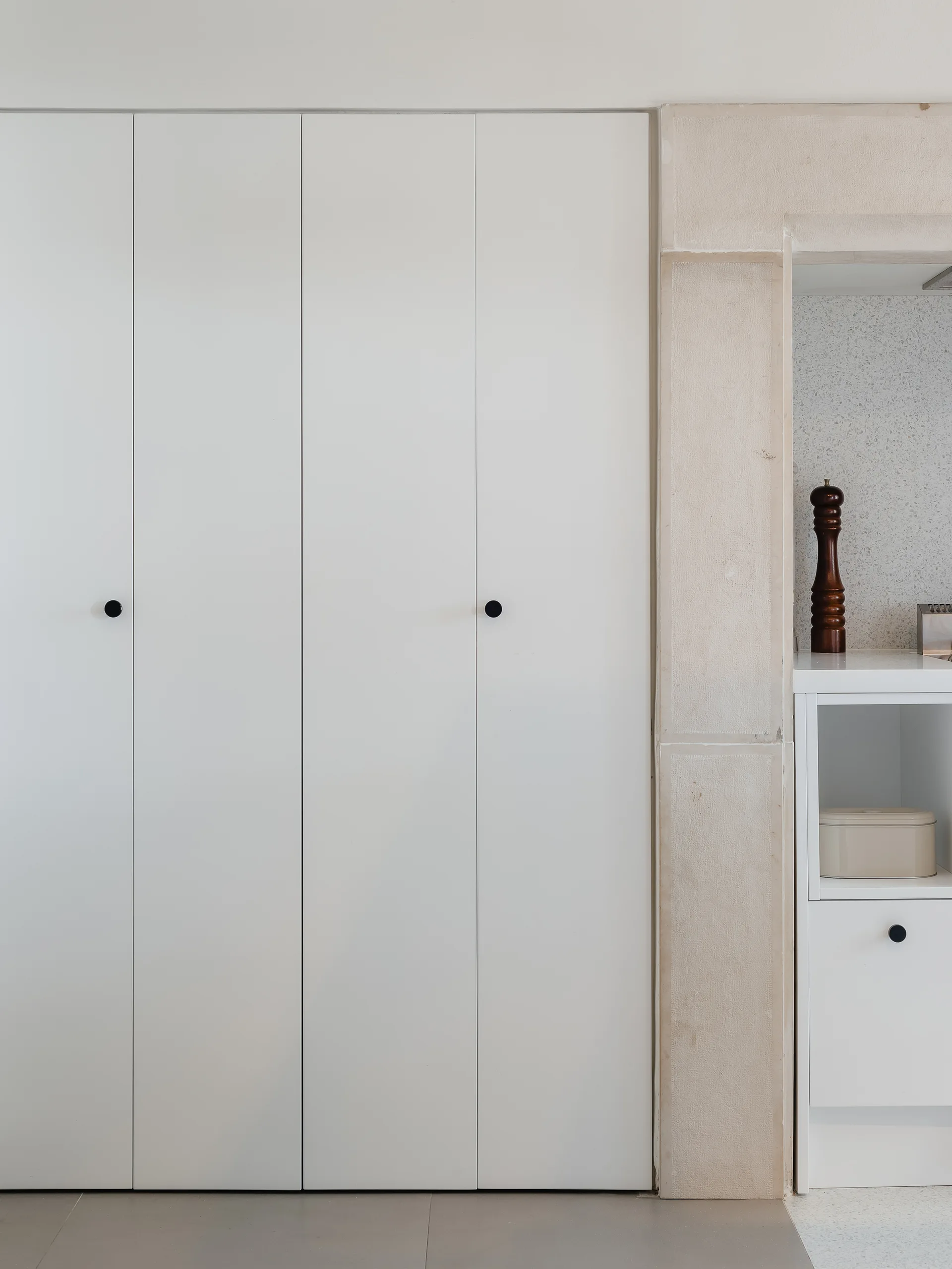 Floor-to-ceiling matte white kitchen cabinets with tiny black round knobs next to a textured beige stone pillar framing an open shelf unit.