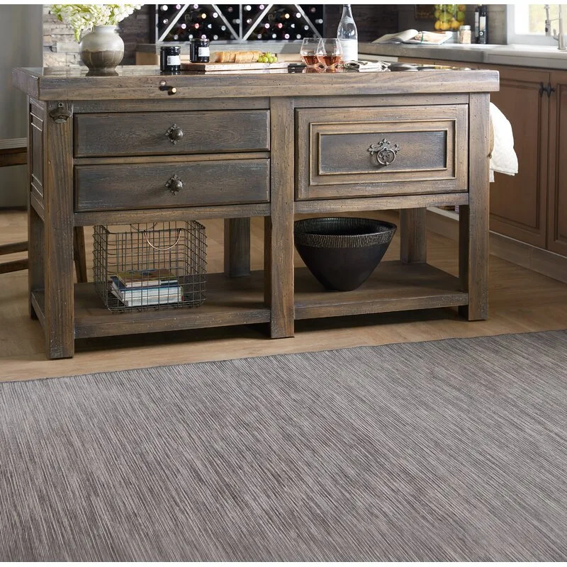 A rustic brown wooden kitchen island featuring drawers with vintage pulls and an open lower shelf rests on a textured grey area rug.