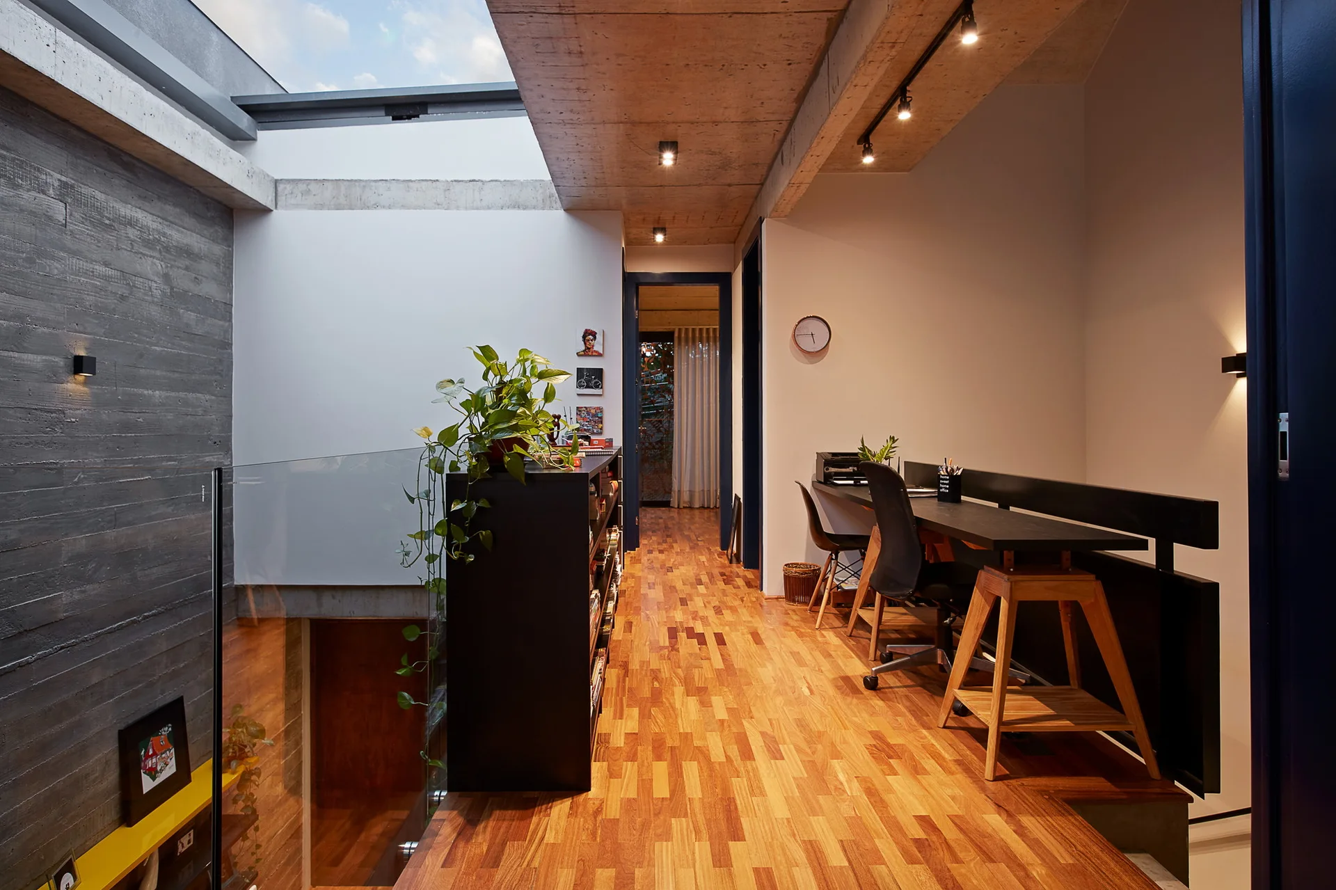 A mezzanine home office features a long black desk, warm wood flooring, a glass railing, and an exposed concrete ceiling with a skylight.