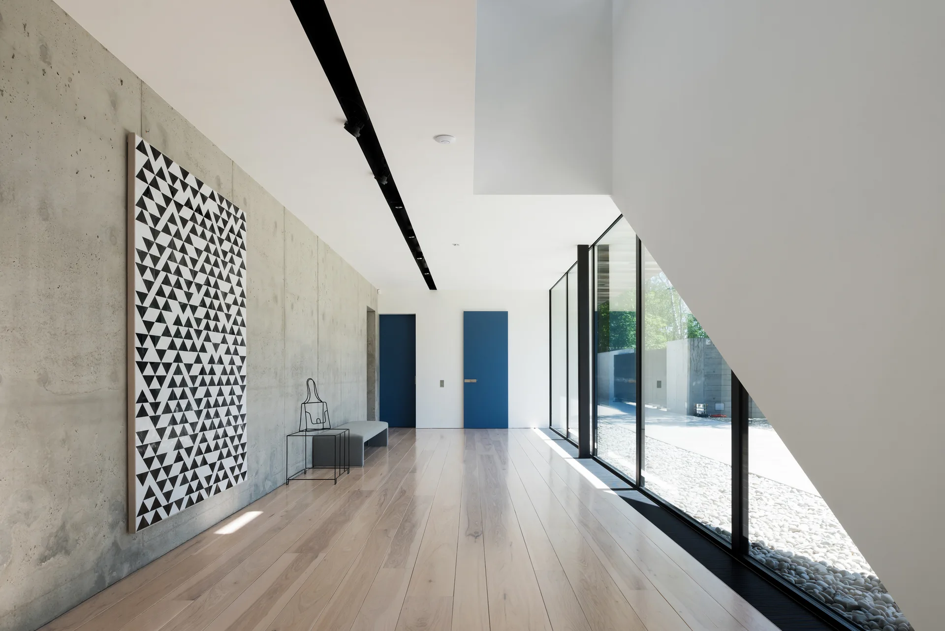 A long hallway features a textured grey concrete wall with black and white art, light wood plank floors, and two blue doors.