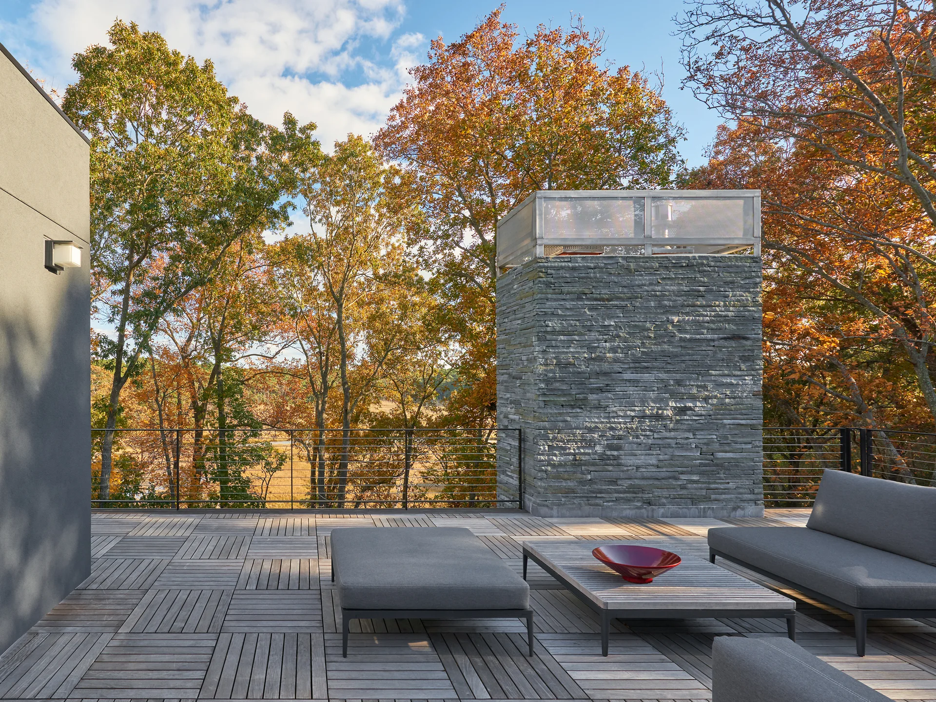 A modern outdoor terrace features grey minimalist lounge furniture, a grid-patterned wood deck, and a tall stacked-stone chimney against autumn trees.