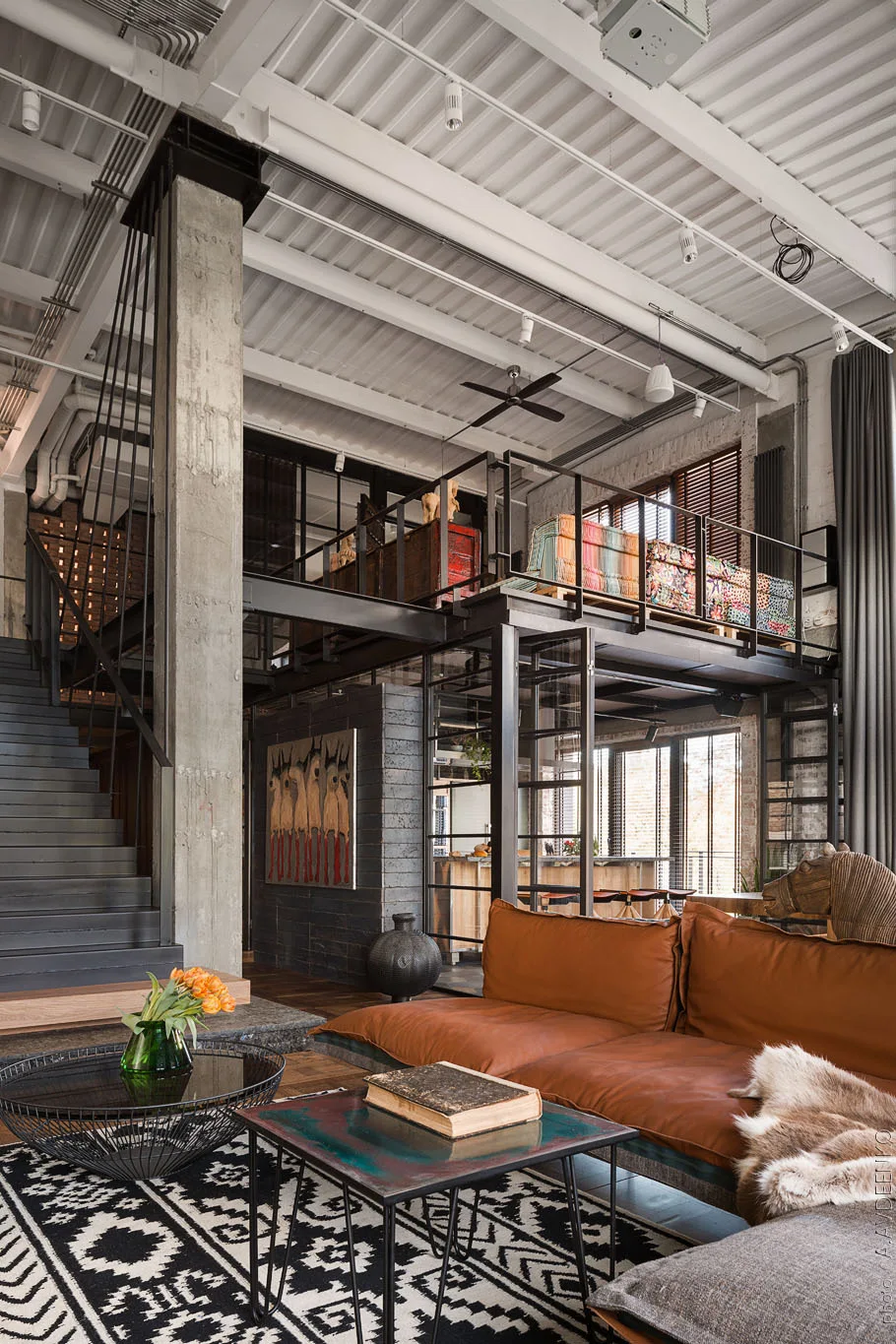 An industrial loft living room with a concrete pillar, a brown leather sofa, a black metal staircase, and an elevated mezzanine level.