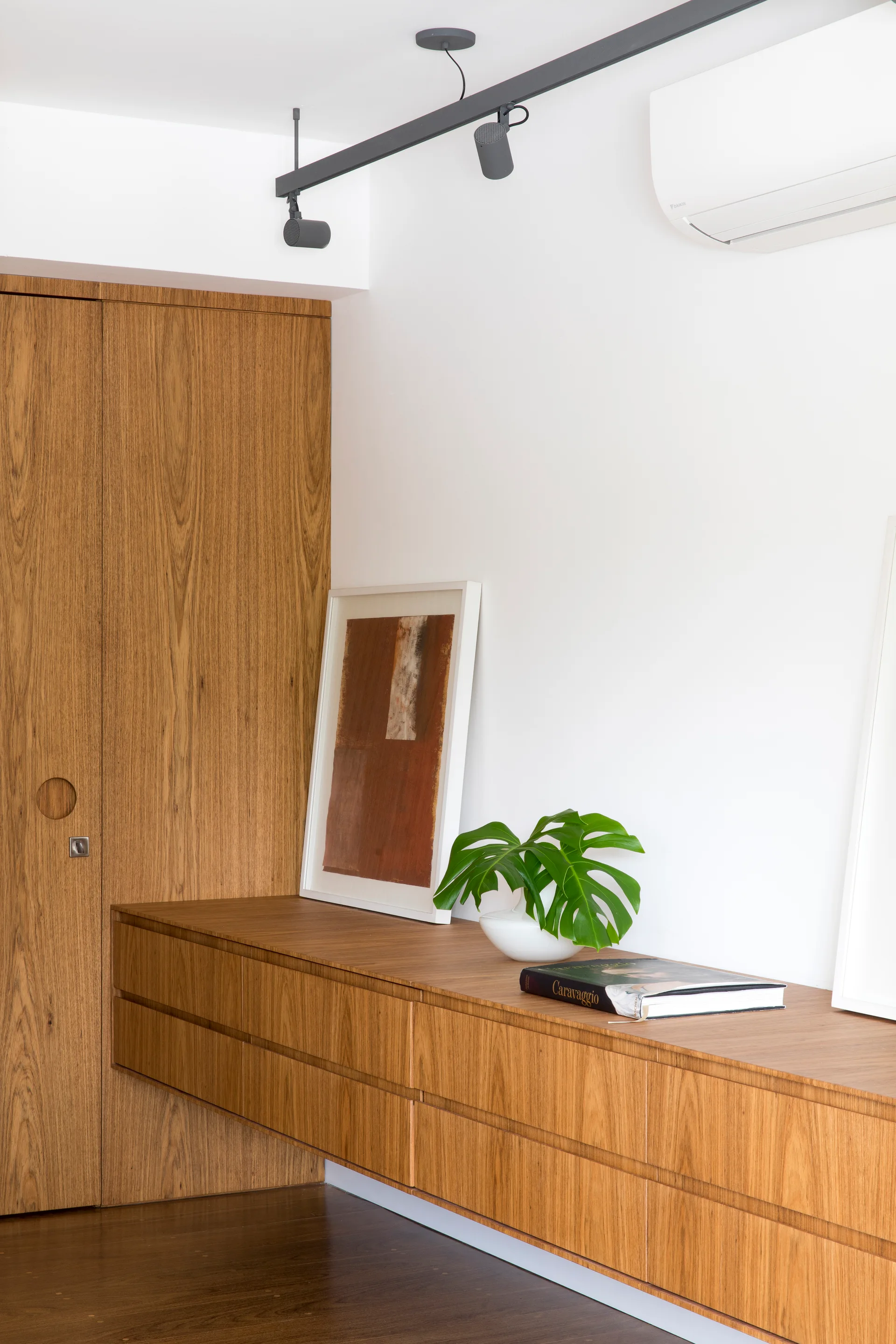 A floating wood credenza and tall built-in closet are positioned against white walls beneath black track lighting and simple decor.