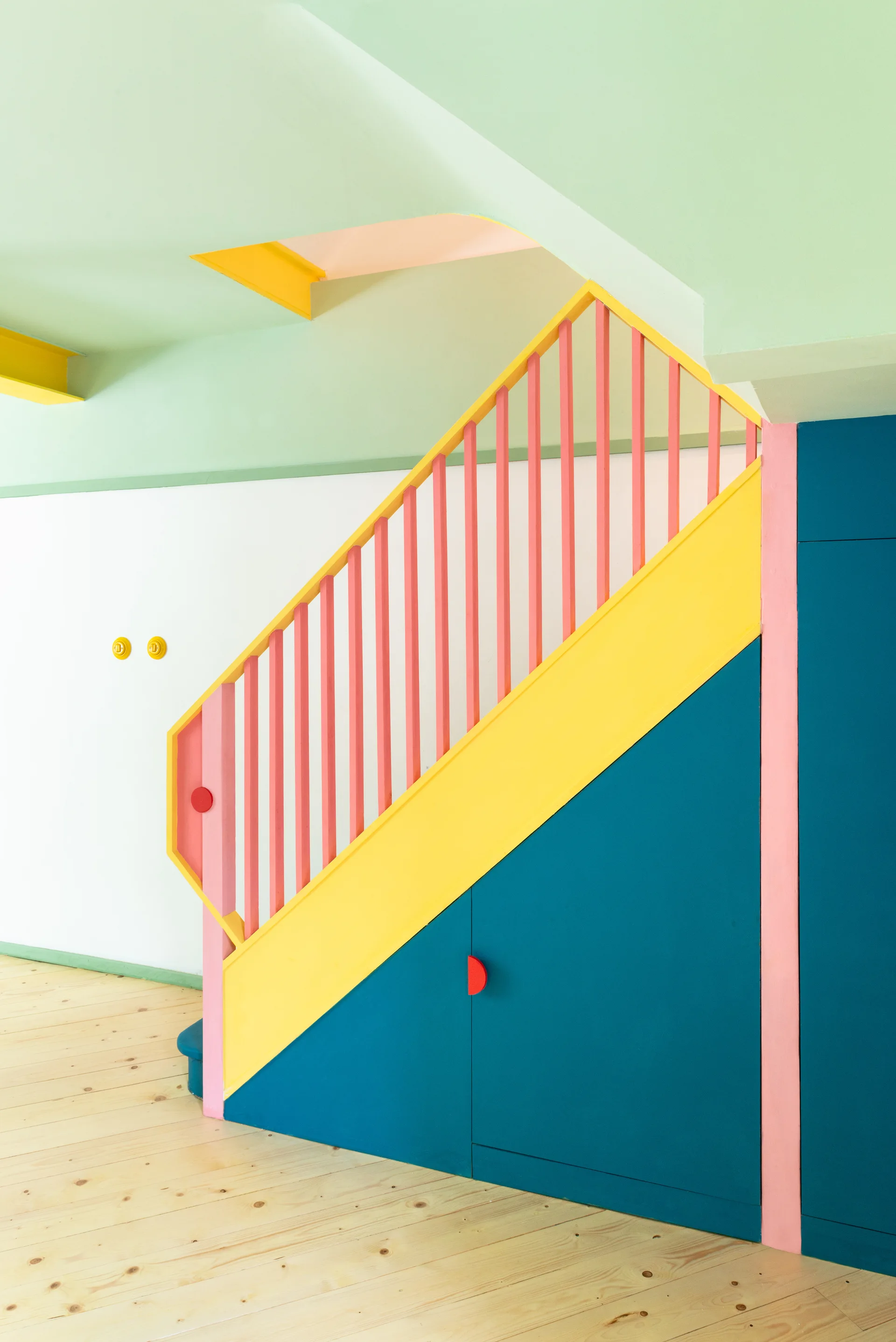 A geometric staircase features pink balusters, a yellow handrail, and blue under-stair storage cabinets over light wood plank flooring.