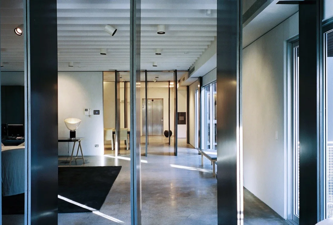 A minimalist hallway featuring dark metal vertical partition columns, a white grid ceiling, polished concrete flooring, and distant elevator doors.