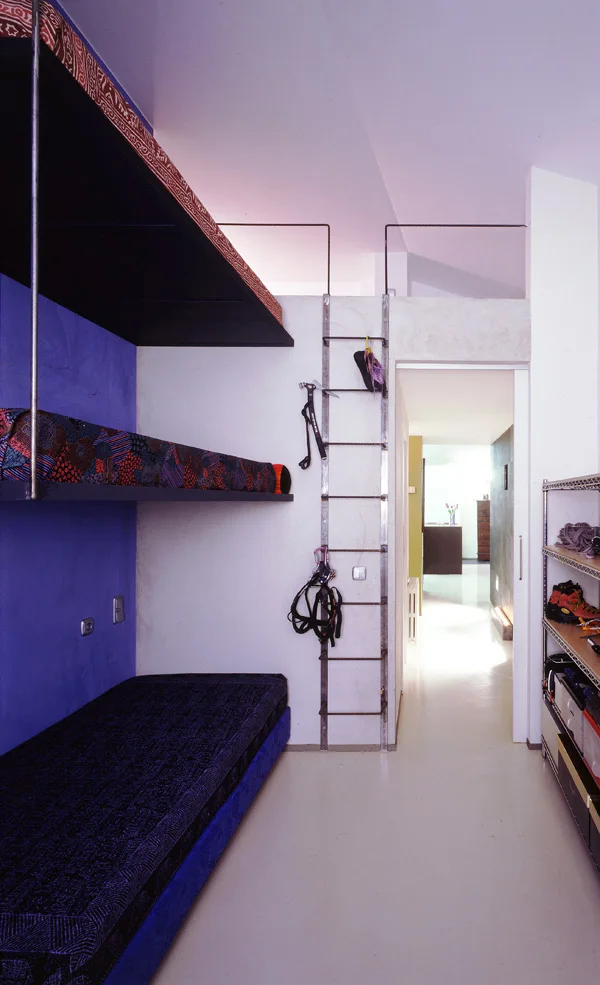 A minimalist bedroom features built-in bunk beds against a blue wall, a steel climbing ladder leading to a loft, and an open doorway.