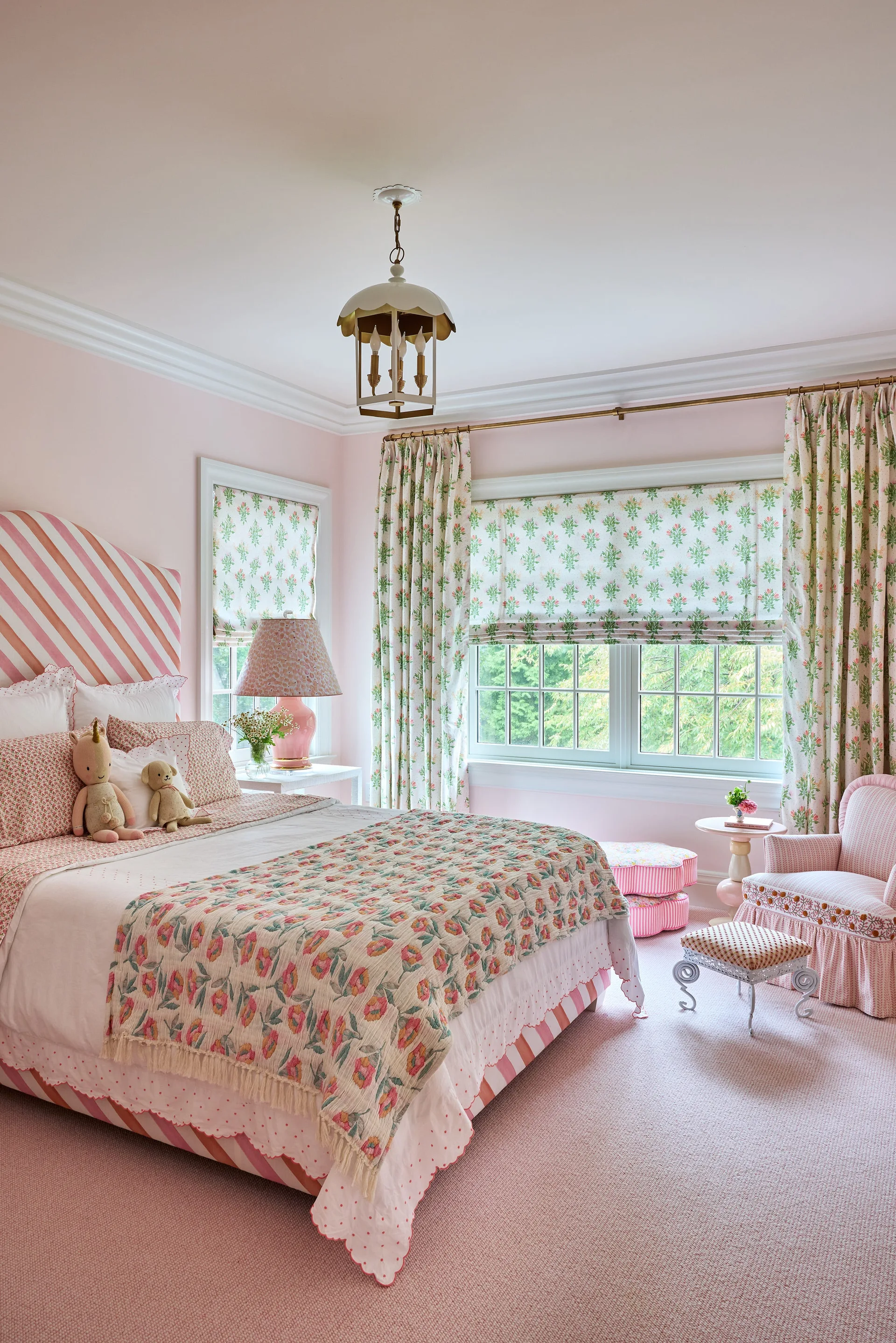 A pink child's bedroom features an upholstered striped headboard, floral patterned quilts, matching drapes, and a ruffled armchair over a pink carpet.
