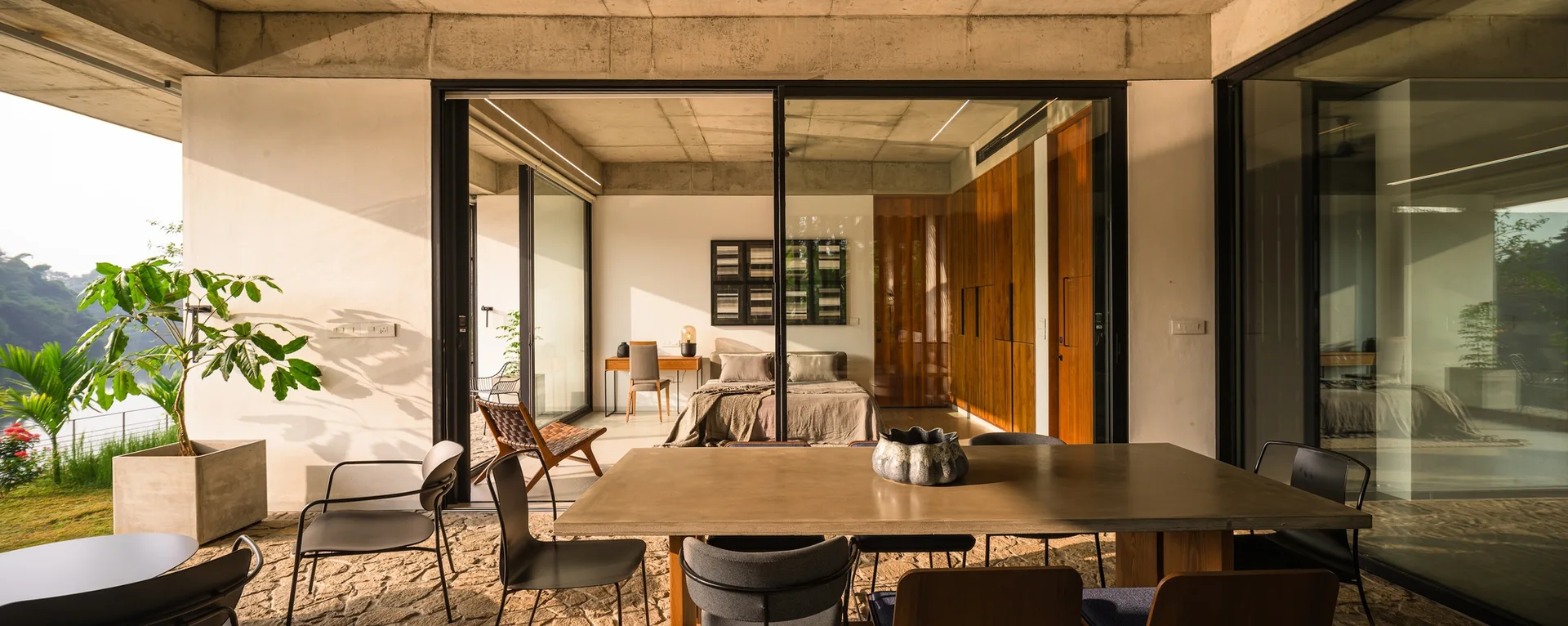 A concrete patio with a dining table transitions through sliding glass doors into a bedroom featuring a minimalist bed and full-height wood cabinetry.