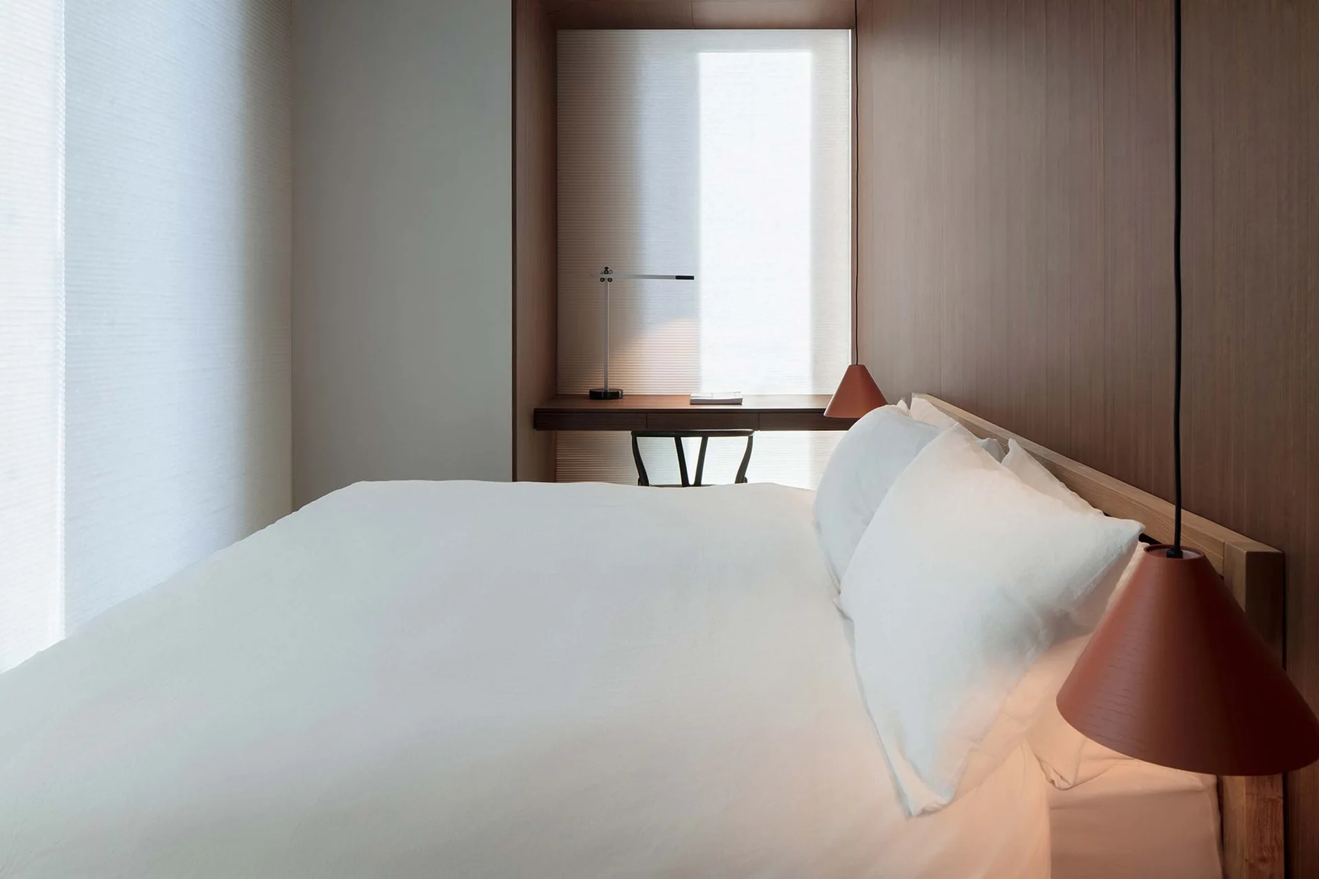 A minimalist bedroom features wood paneled walls, a bed with white linens, terracotta pendant lights, and a built-in wooden desk in a window alcove.