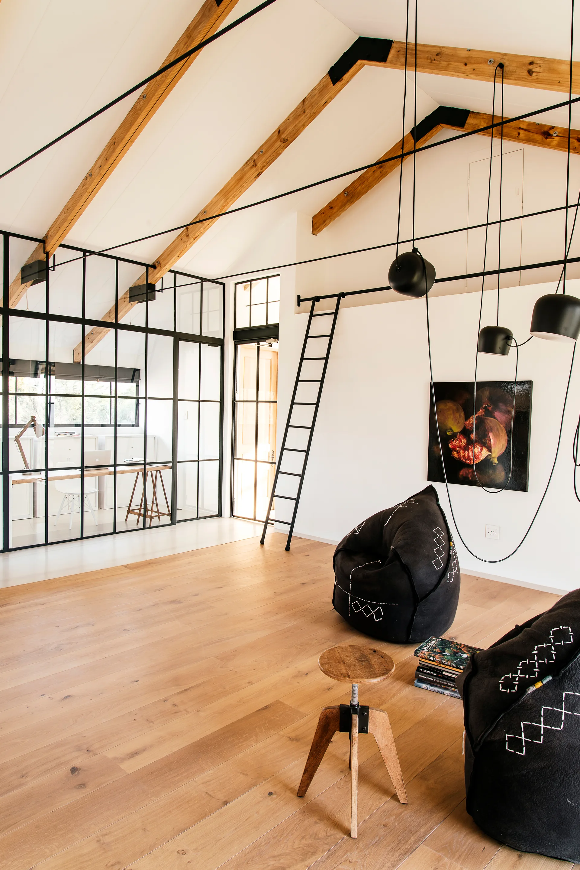 A loft space featuring a vaulted ceiling with timber beams, a black-framed glass partition, and black bean bag chairs on light wood floors.