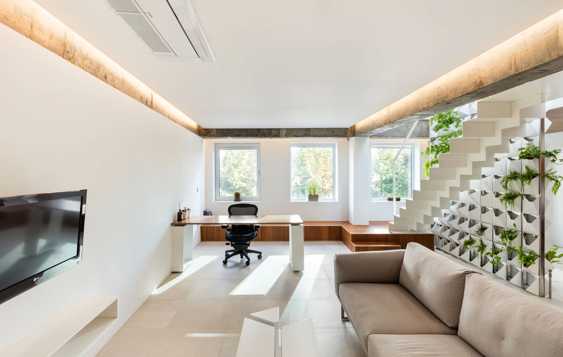A modern open-plan living room and workspace featuring a beige sofa, a wood desk under windows, and a white staircase integrated with green planters.