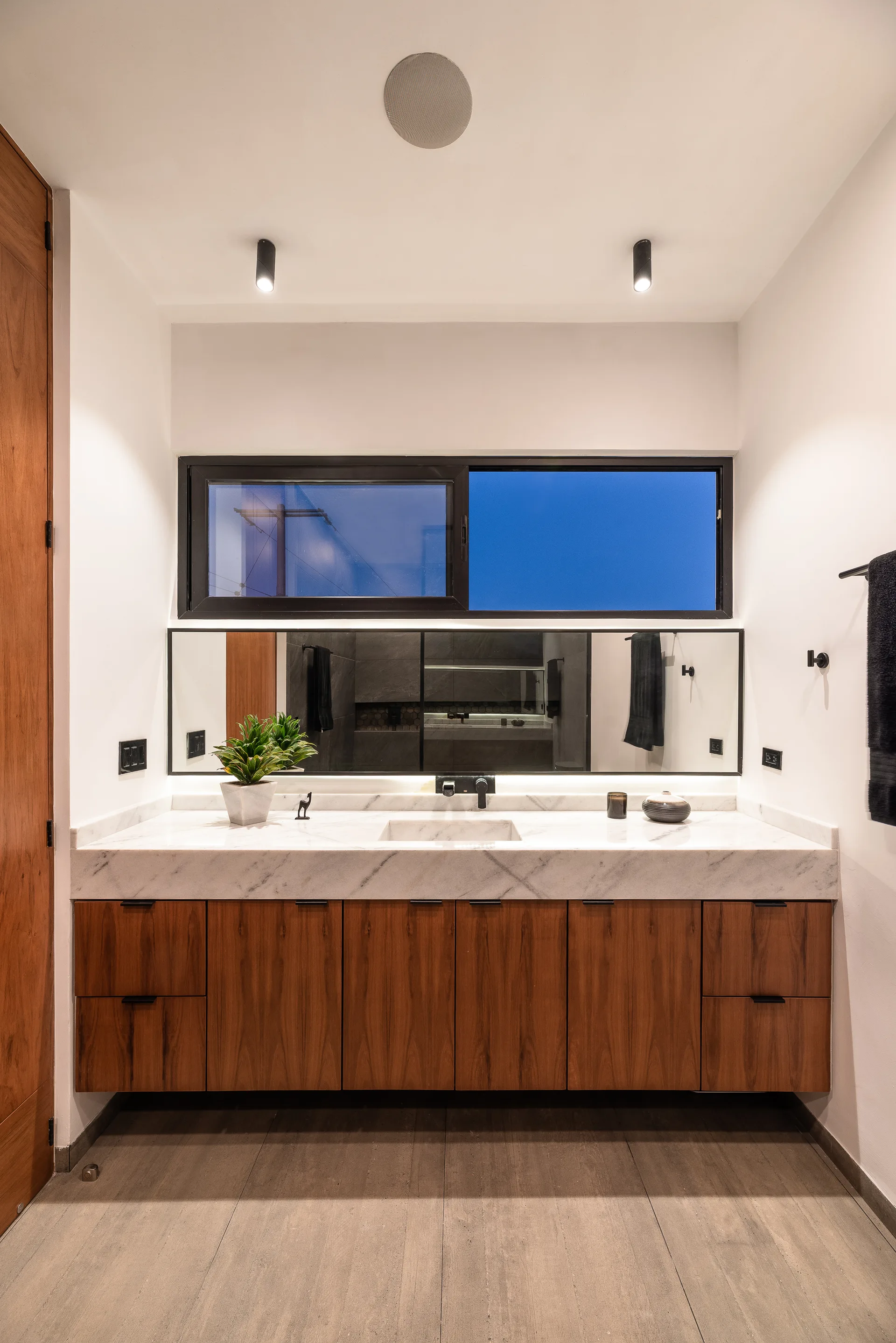 A floating wood bathroom vanity features a white marble countertop with an integrated sink beneath a wide horizontal mirror and window.