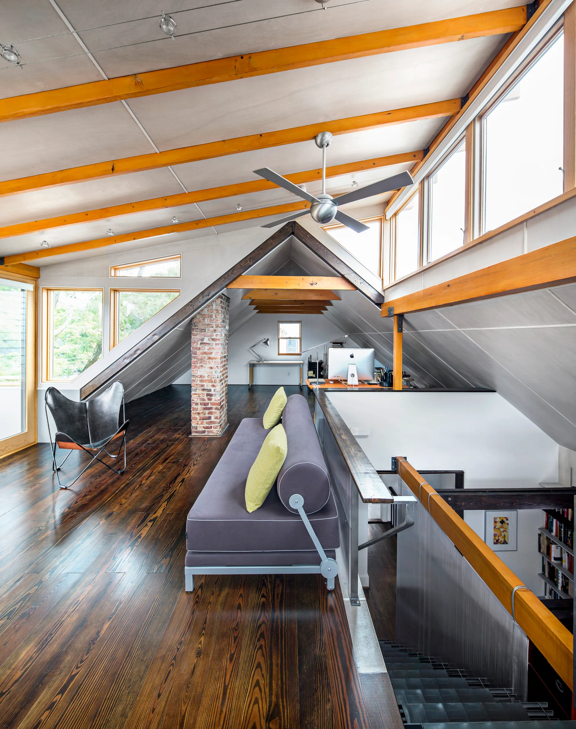 An open mezzanine loft features dark wood flooring, sloped white ceilings with exposed timber beams, a grey sofa, and a brick column.