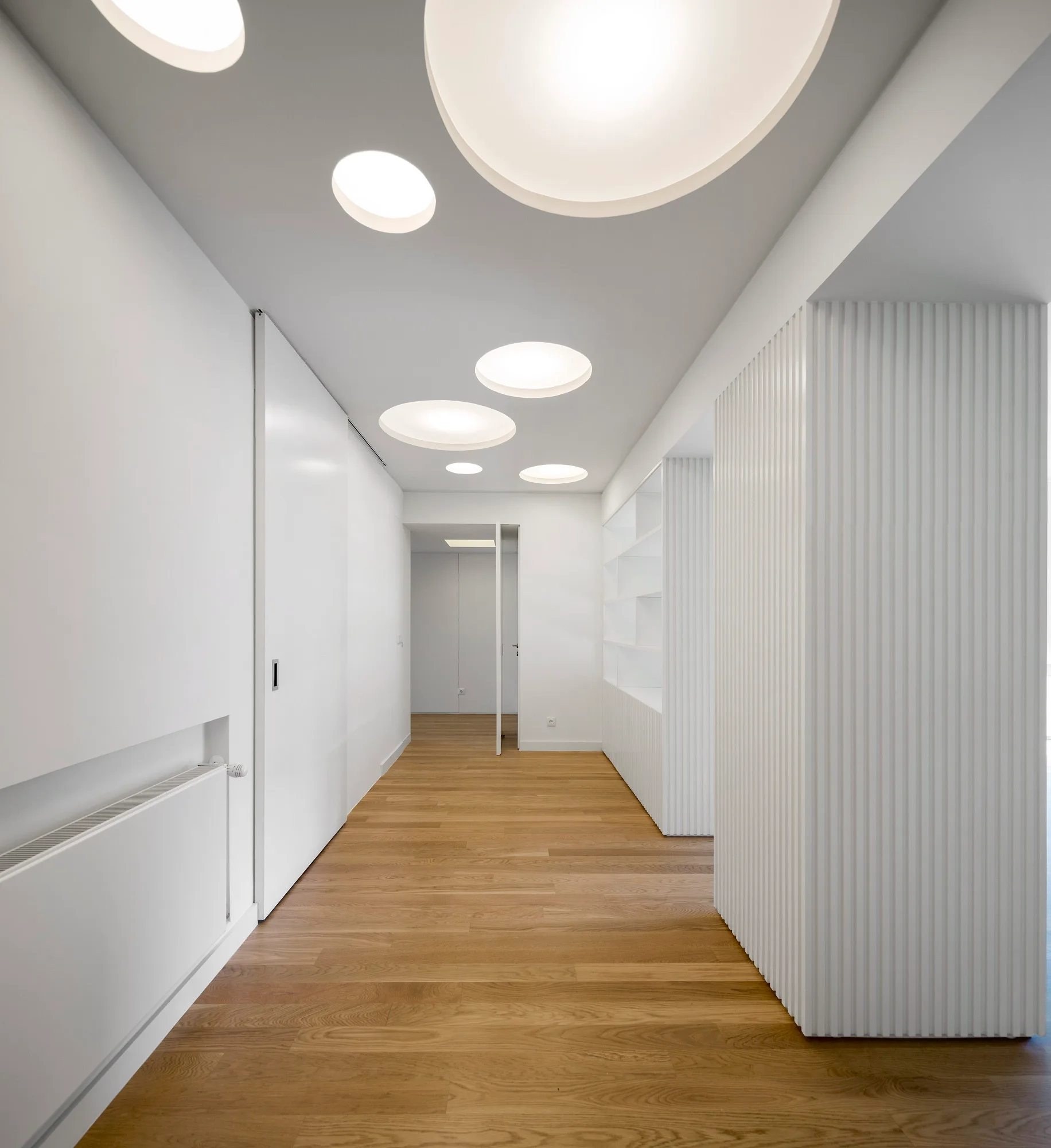 A minimalist white hallway features wood flooring, vertical ribbed wall paneling, built-in shelving, and large circular recessed ceiling lights.
