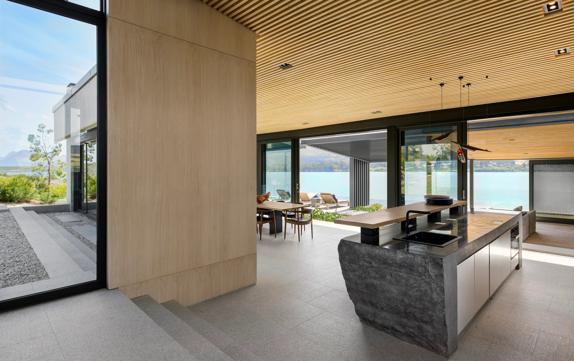A modern kitchen featuring a textured rough-cut stone island, linear wood slat ceiling, and large glass doors looking out onto a lake.
