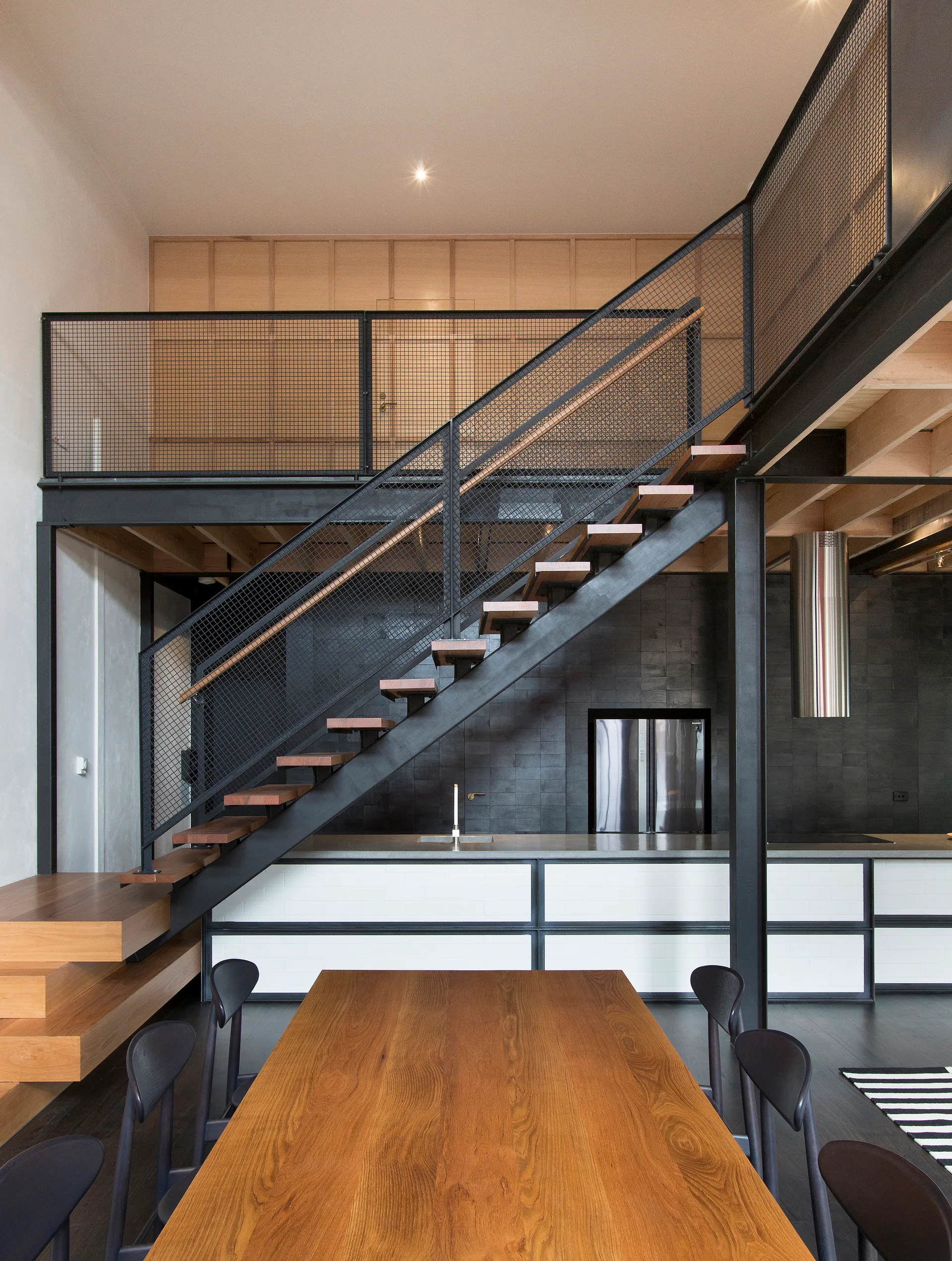 A black steel staircase with wood treads rises over an industrial kitchen featuring white base cabinets and black tiled walls.