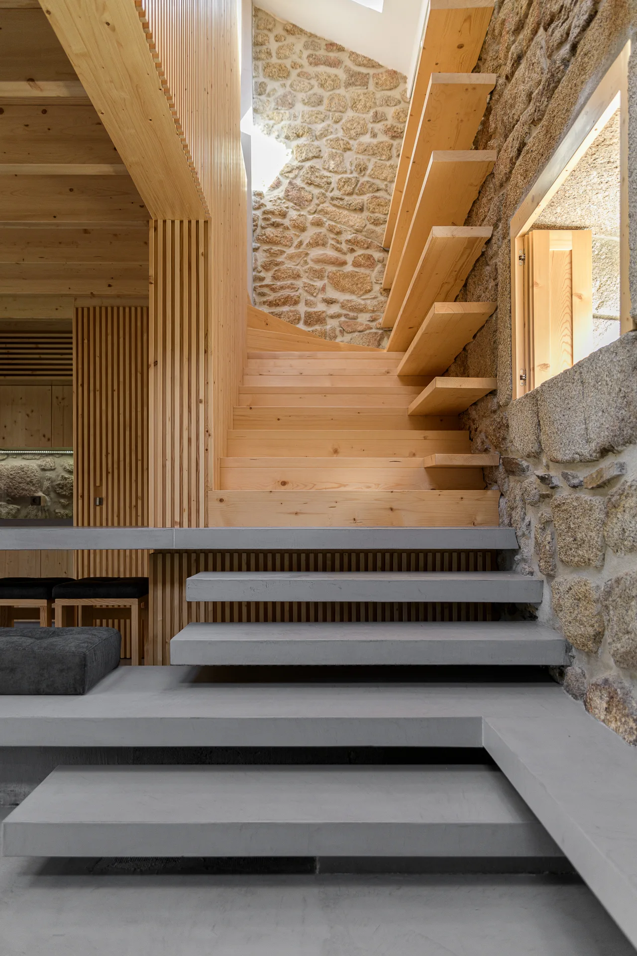 An interior staircase features floating wooden steps set against an exposed stone wall, leading up from tiered grey concrete platform steps.