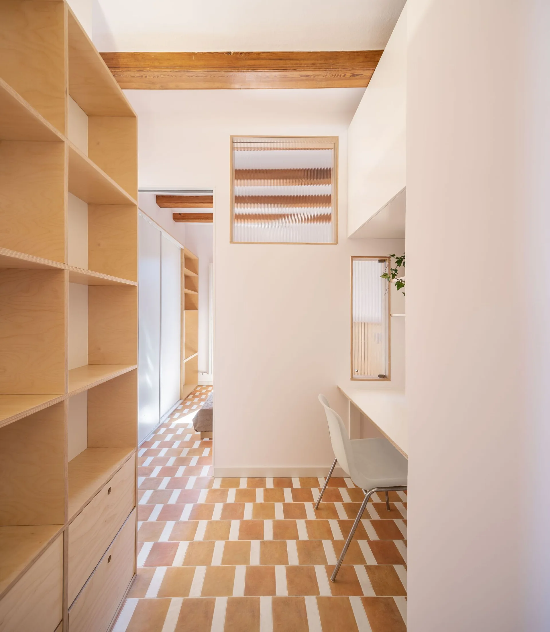 A minimalist study nook features built-in plywood shelving, a white desk, reeded glass wall inserts, and geometric terracotta tile flooring.
