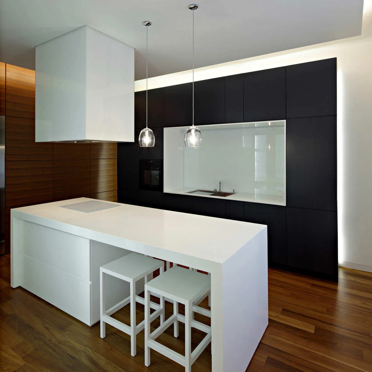 A modern minimalist kitchen features matte black cabinetry, a white island with stools, a blocky white range hood, and warm wood flooring.