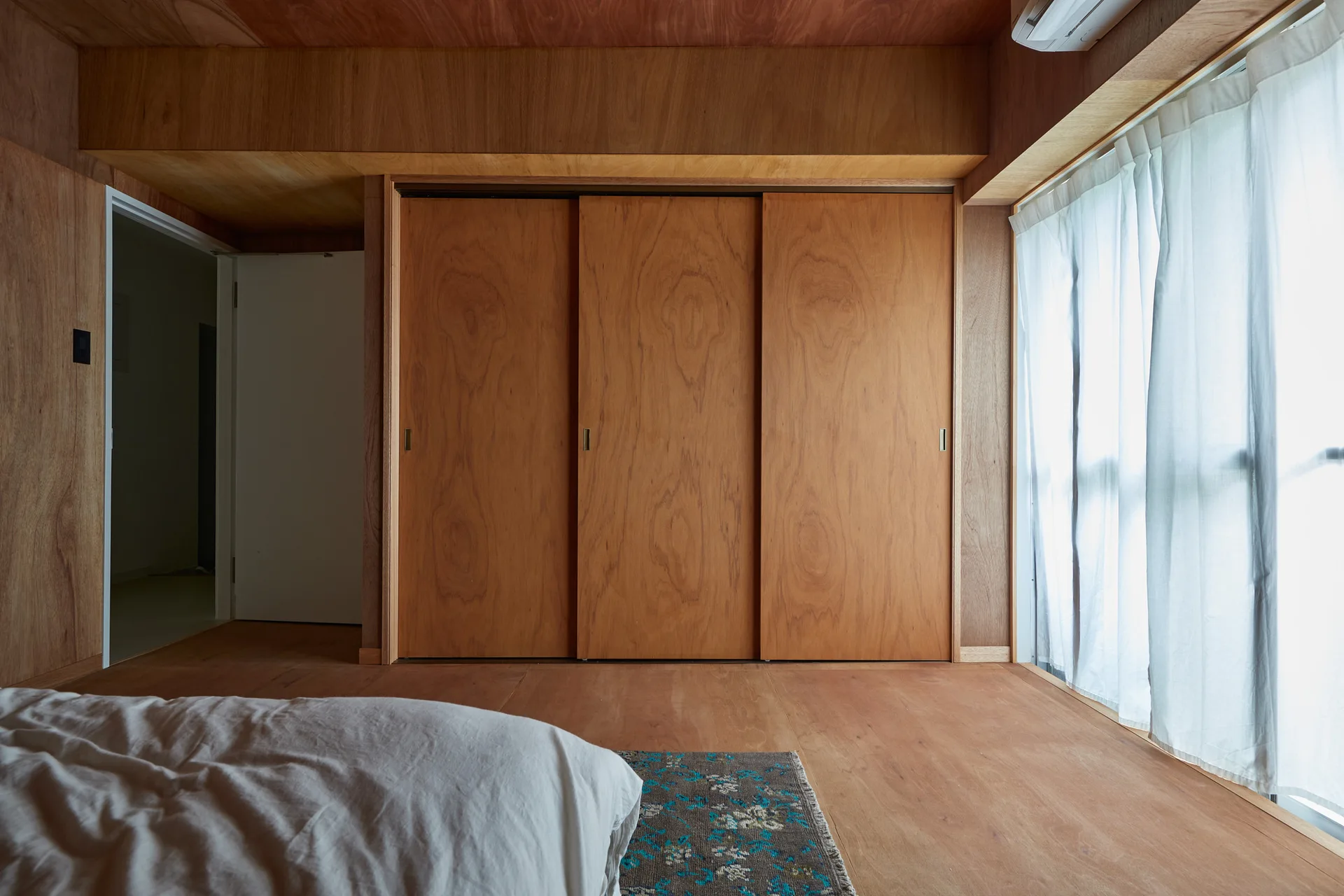 A minimalist bedroom features warm plywood paneling on the walls, ceiling, and floor, with large sliding wardrobe doors and a sheer curtained window.