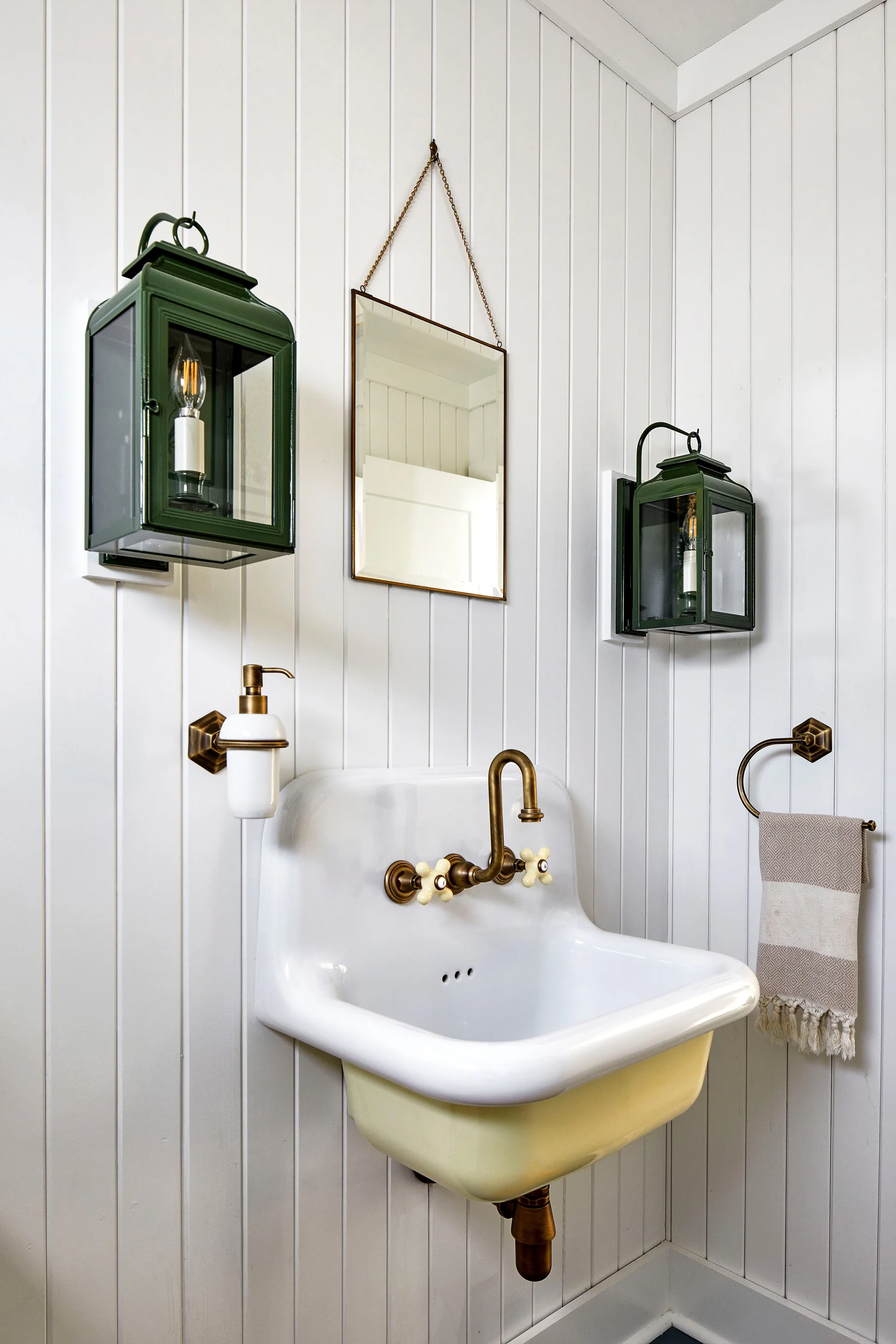 A white wall-mounted utility sink with brass fixtures features two green lantern sconces and a small hanging mirror on a white beadboard wall.