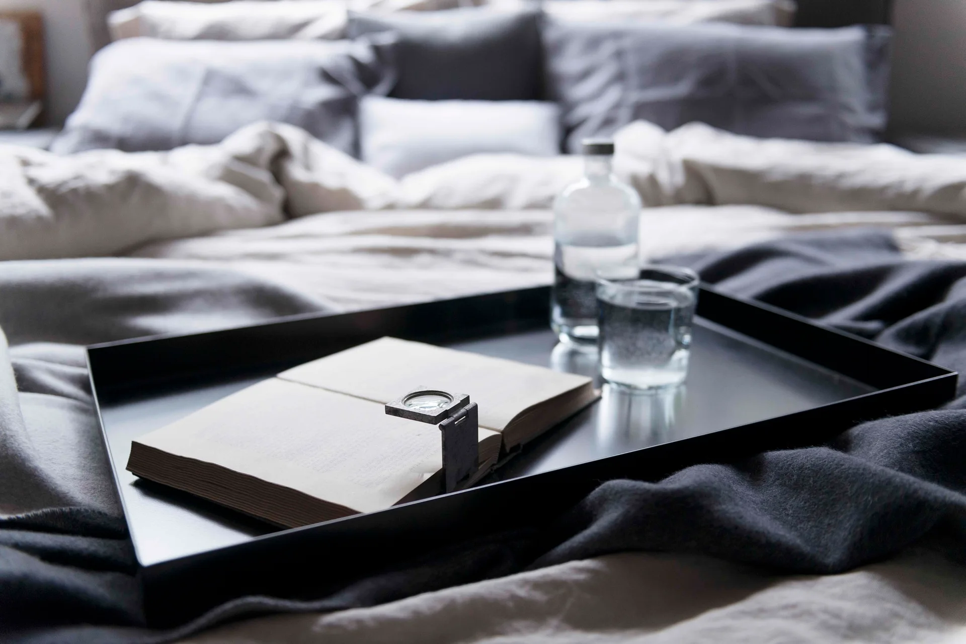 A close-up of a black metal tray on ruffled grey bedding, holding an open book with a magnifying glass, a water bottle, and a glass of water.