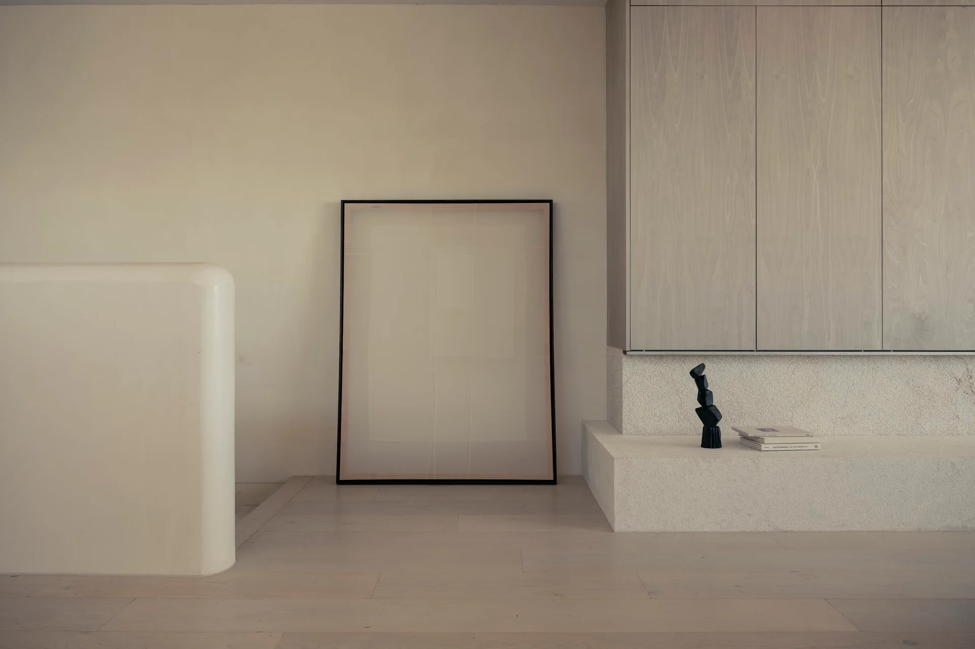 A minimalist room features a floating light wood cabinet over a textured stone platform, with a large framed artwork leaning against the wall.