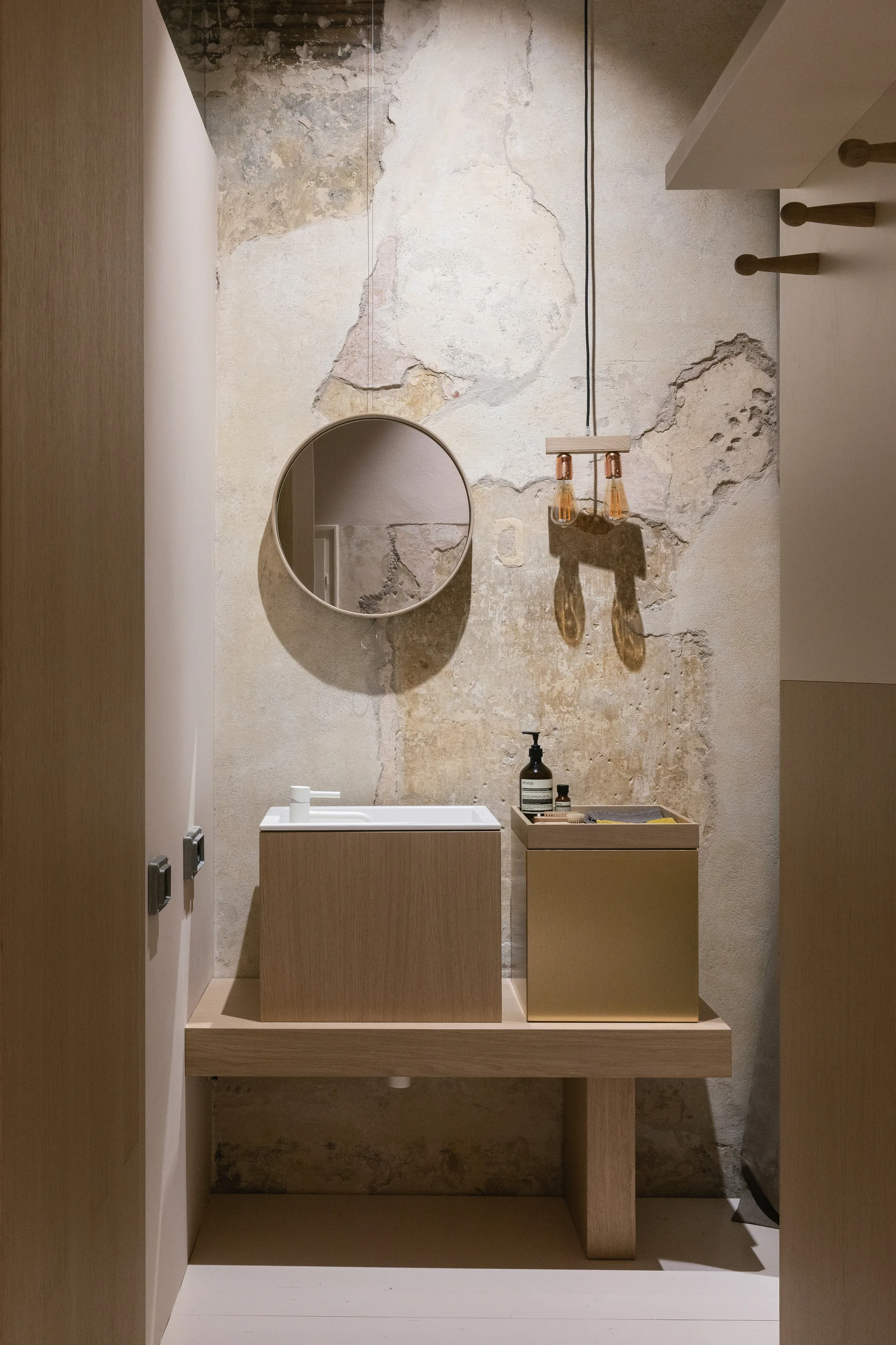 A minimalist wooden bathroom vanity with a white basin stands against a weathered, distressed plaster wall beneath a round mirror and hanging pendant light.
