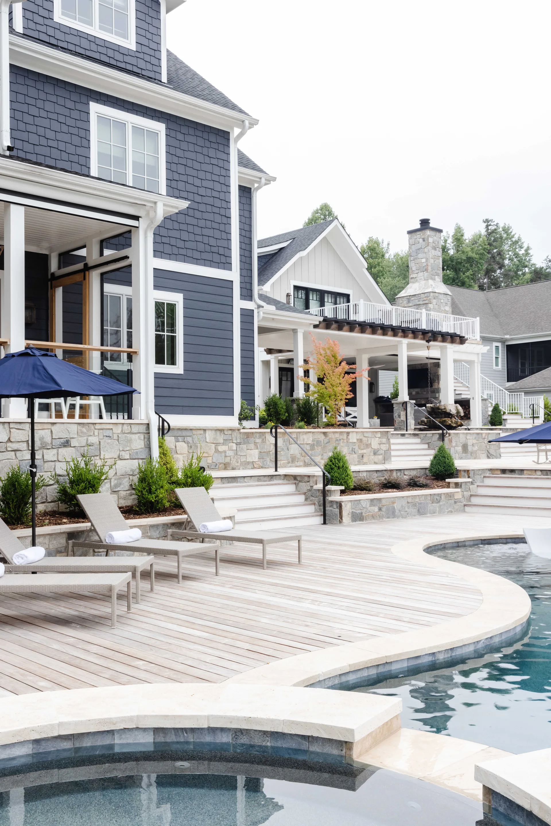 A backyard swimming pool features a weathered wood deck, wicker lounge chairs, tiered stone retaining walls, and a house with navy shingle siding.