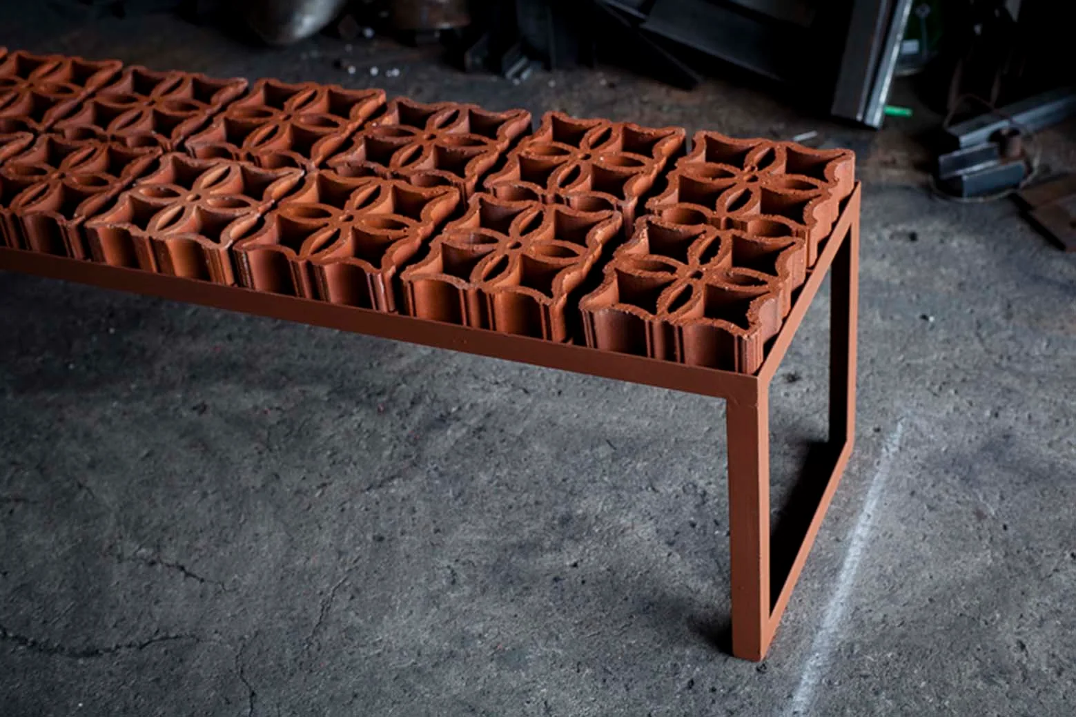 A long metal bench featuring a row of decorative terracotta screen blocks sits on a concrete floor in a workshop.