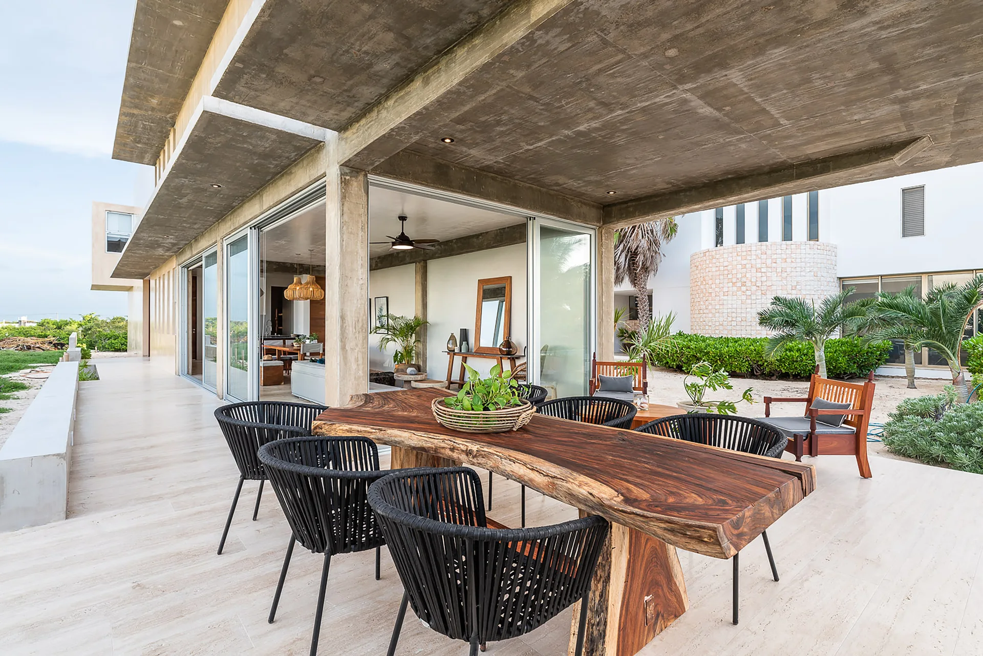 A modern covered patio features a live-edge wood dining table paired with black woven chairs under an exposed concrete ceiling.