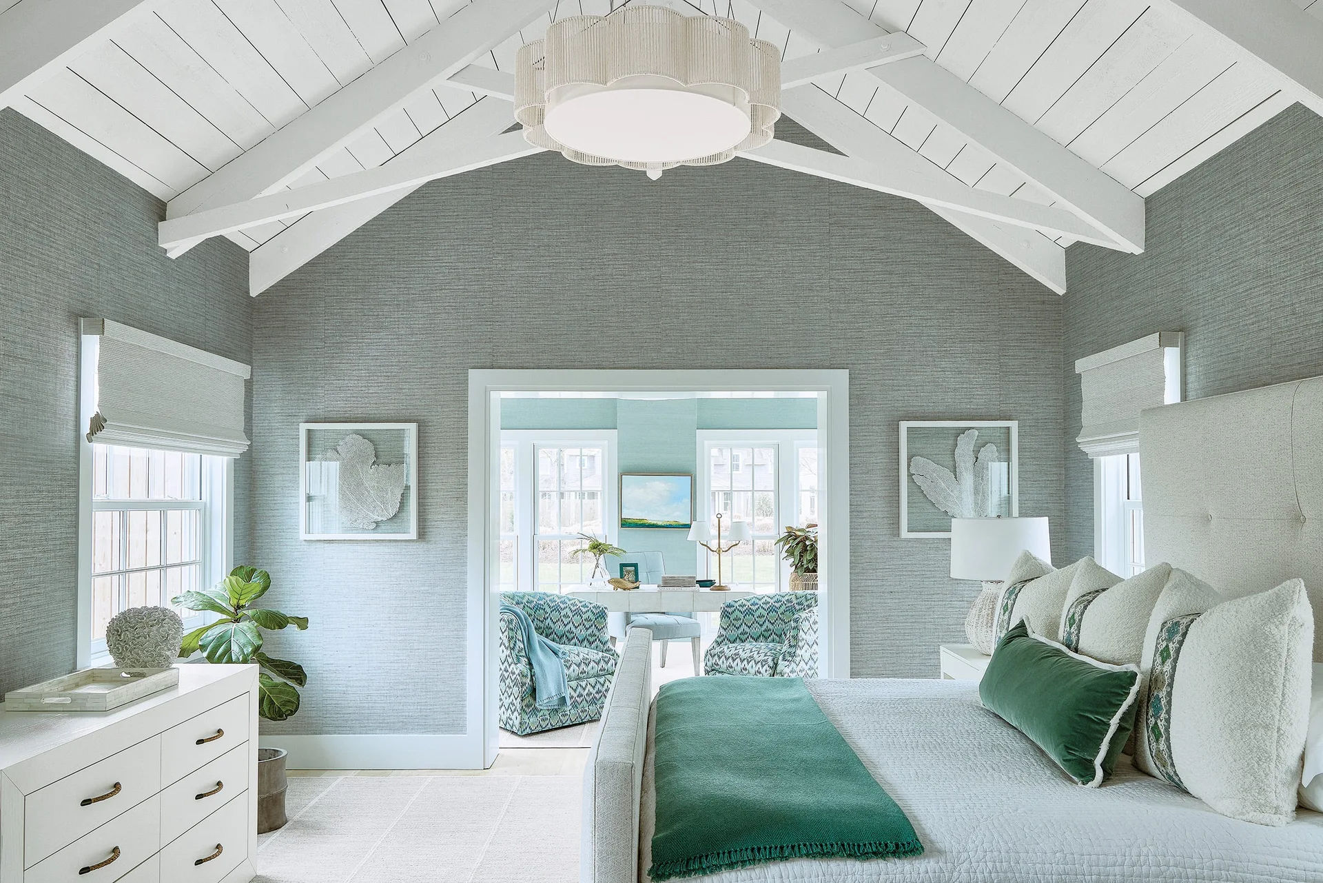 A bedroom with gray textured wallpaper, a white vaulted ceiling with wooden beams, and an open doorway leading to a bright sitting room.