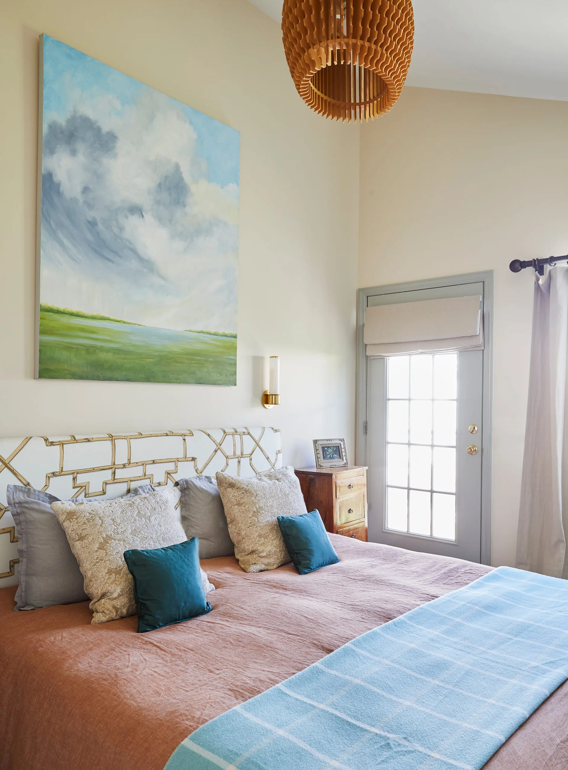 A bedroom features a bed with terracotta-toned linens, an upholstered trellis pattern headboard, a large cloud painting, and a slatted wood pendant light.