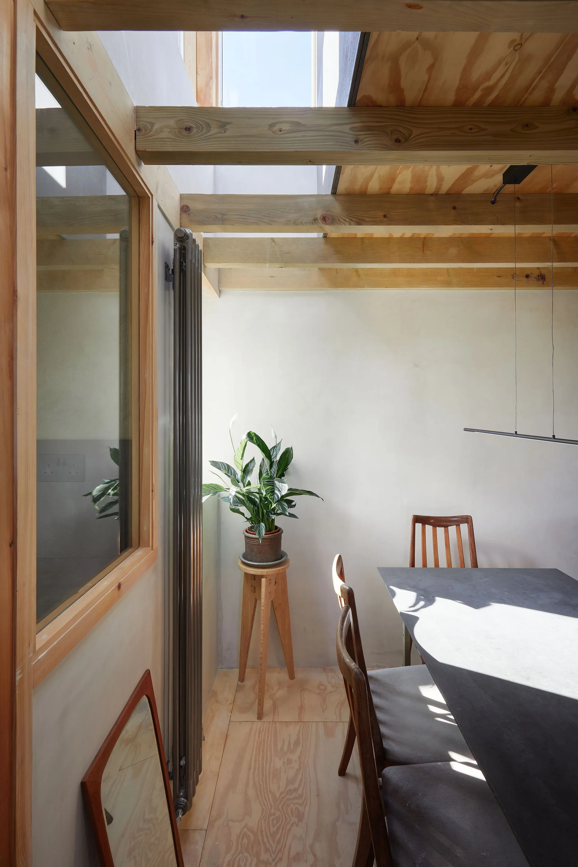 A minimalist dining area featuring a dark gray table, exposed wood ceiling beams, a skylight, and a potted plant on a stool.