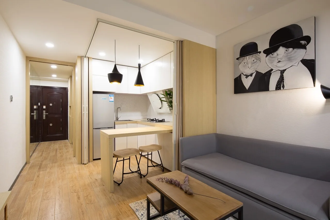 A compact apartment interior featuring a grey sofa, a small wooden breakfast bar with two stools, and a kitchen separated by a folding wood partition.