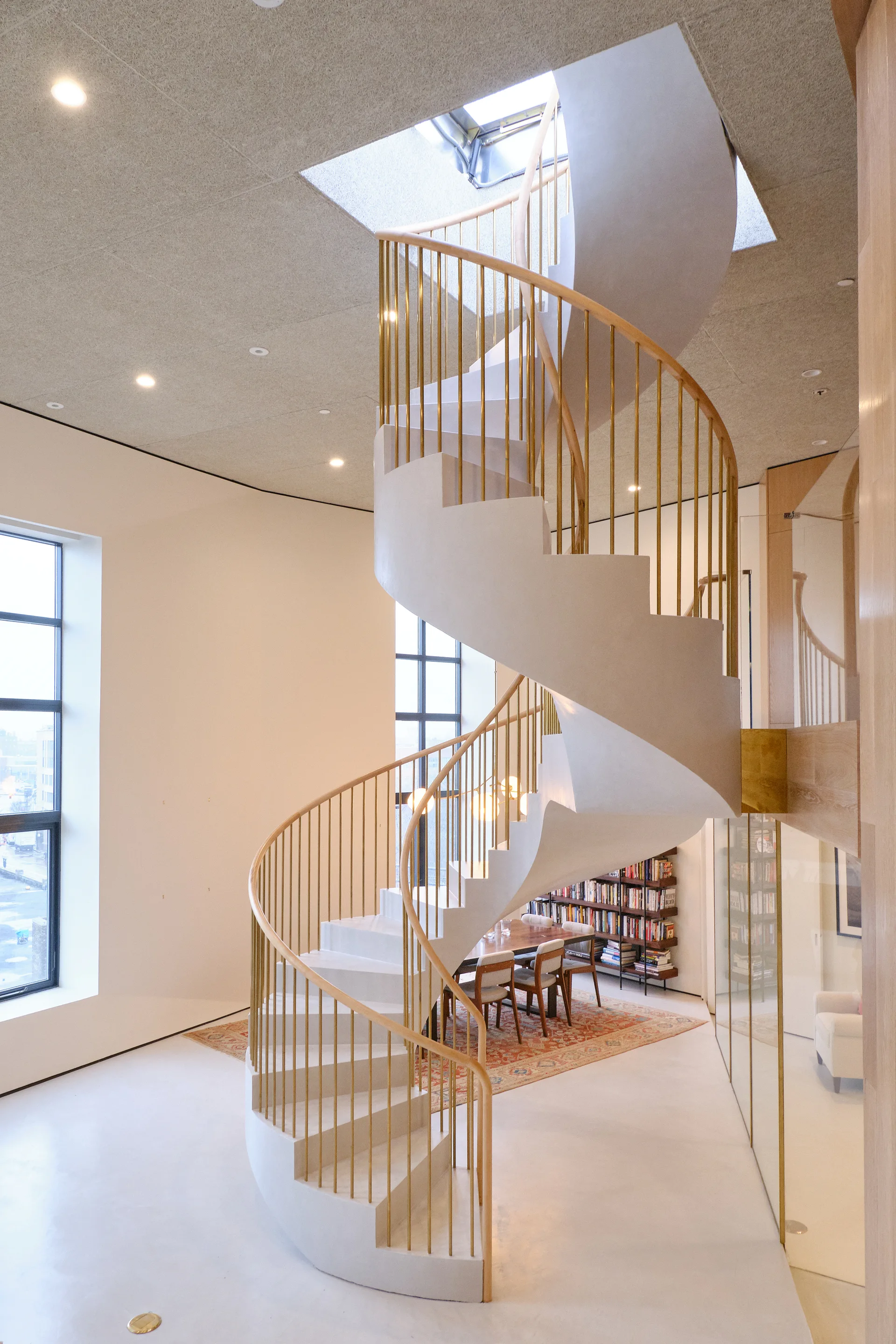 A sculptural white spiral staircase with brass railings rises through a square skylight in a bright room with a dining area.