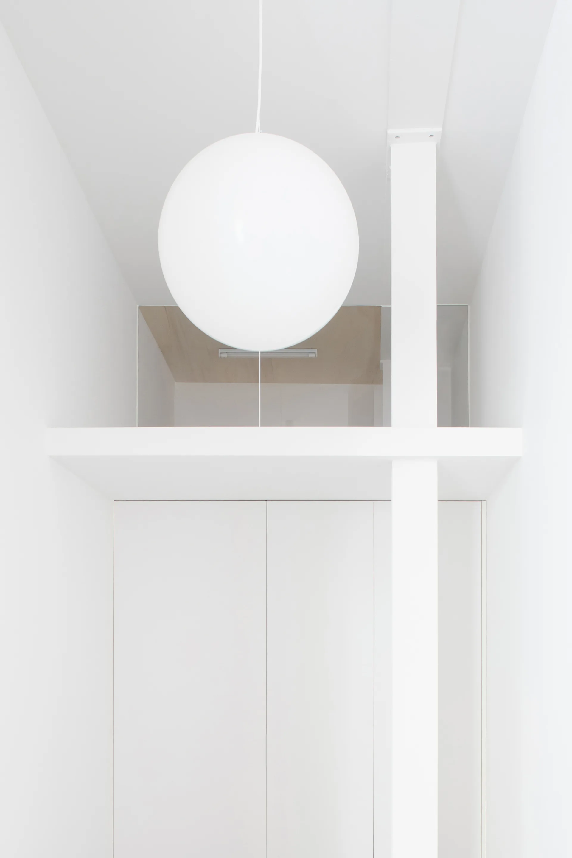 A large white spherical pendant light hangs in a minimalist all-white space above flat-panel cabinetry and a structural column.