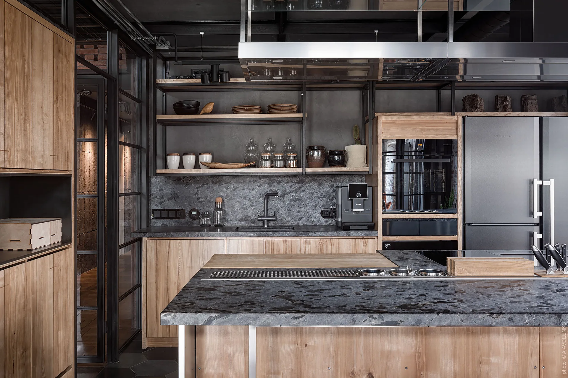 An industrial kitchen features light wood cabinets, dark granite countertops, open metal shelving, and a black-framed glass wall partition.
