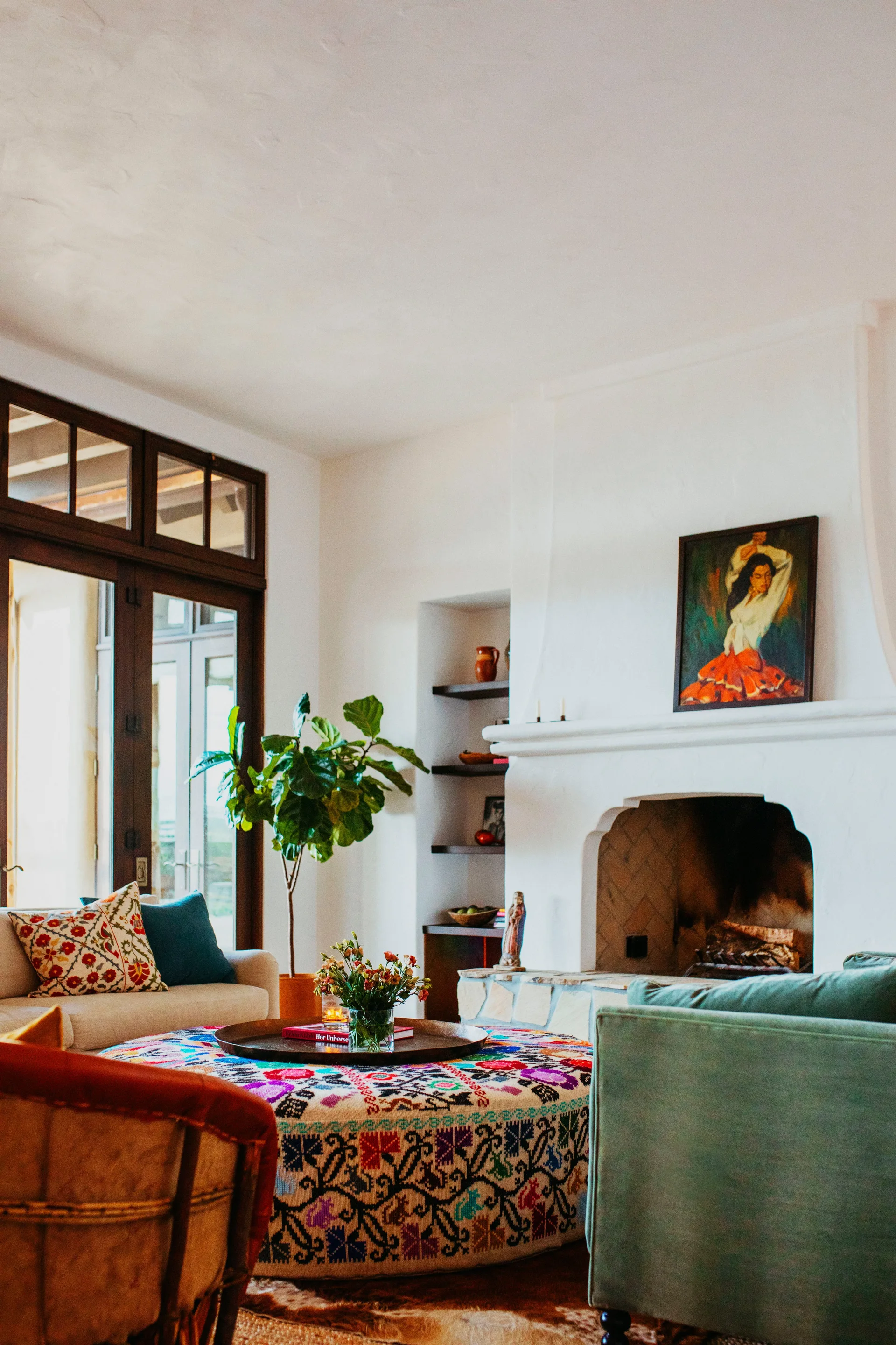 A living room features a white plaster fireplace, a large round embroidered ottoman, and a fiddle leaf fig plant near dark wood glass doors.