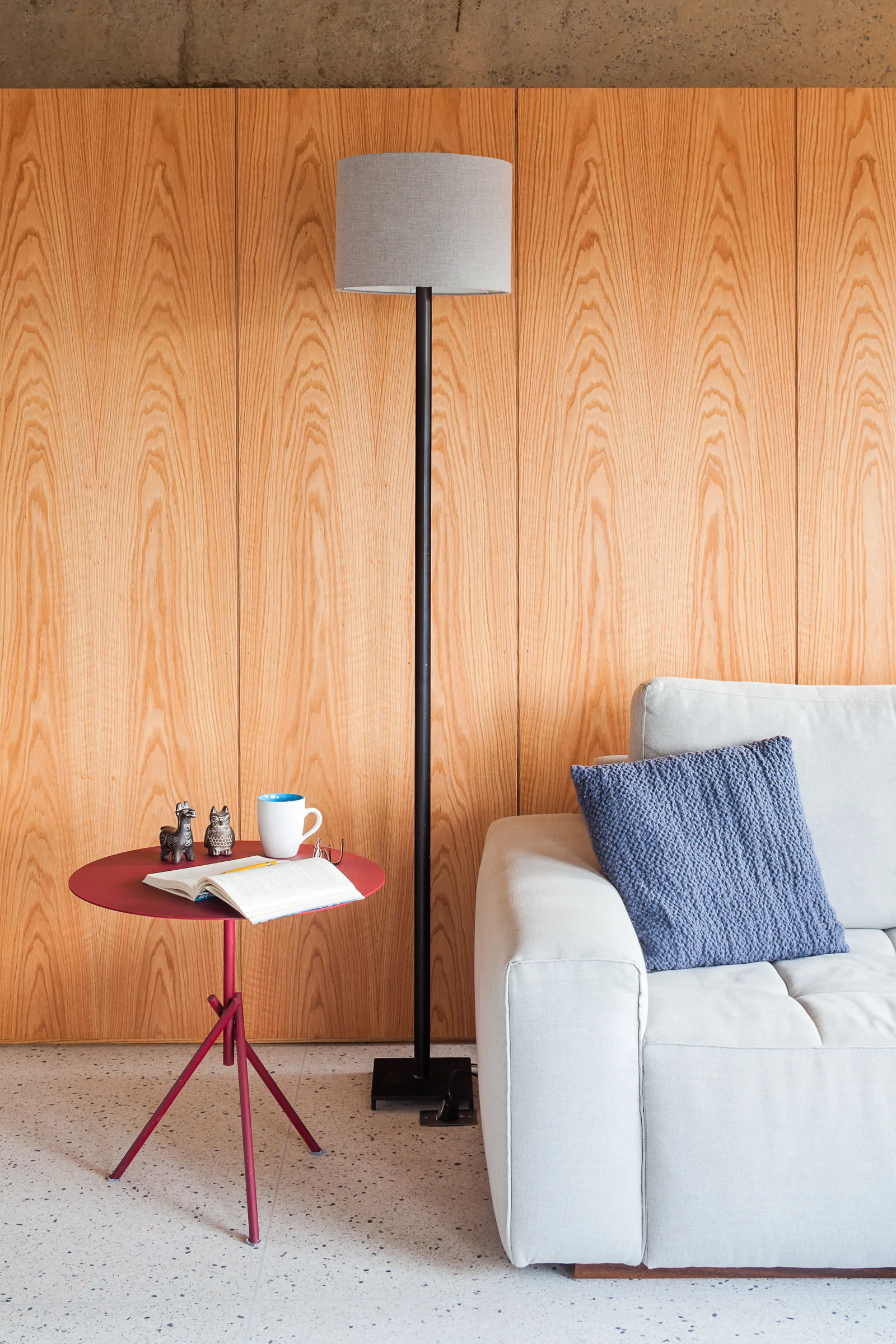 A light grey sofa sits next to a red metal side table and black floor lamp against a vertical wood paneled wall on a terrazzo floor.